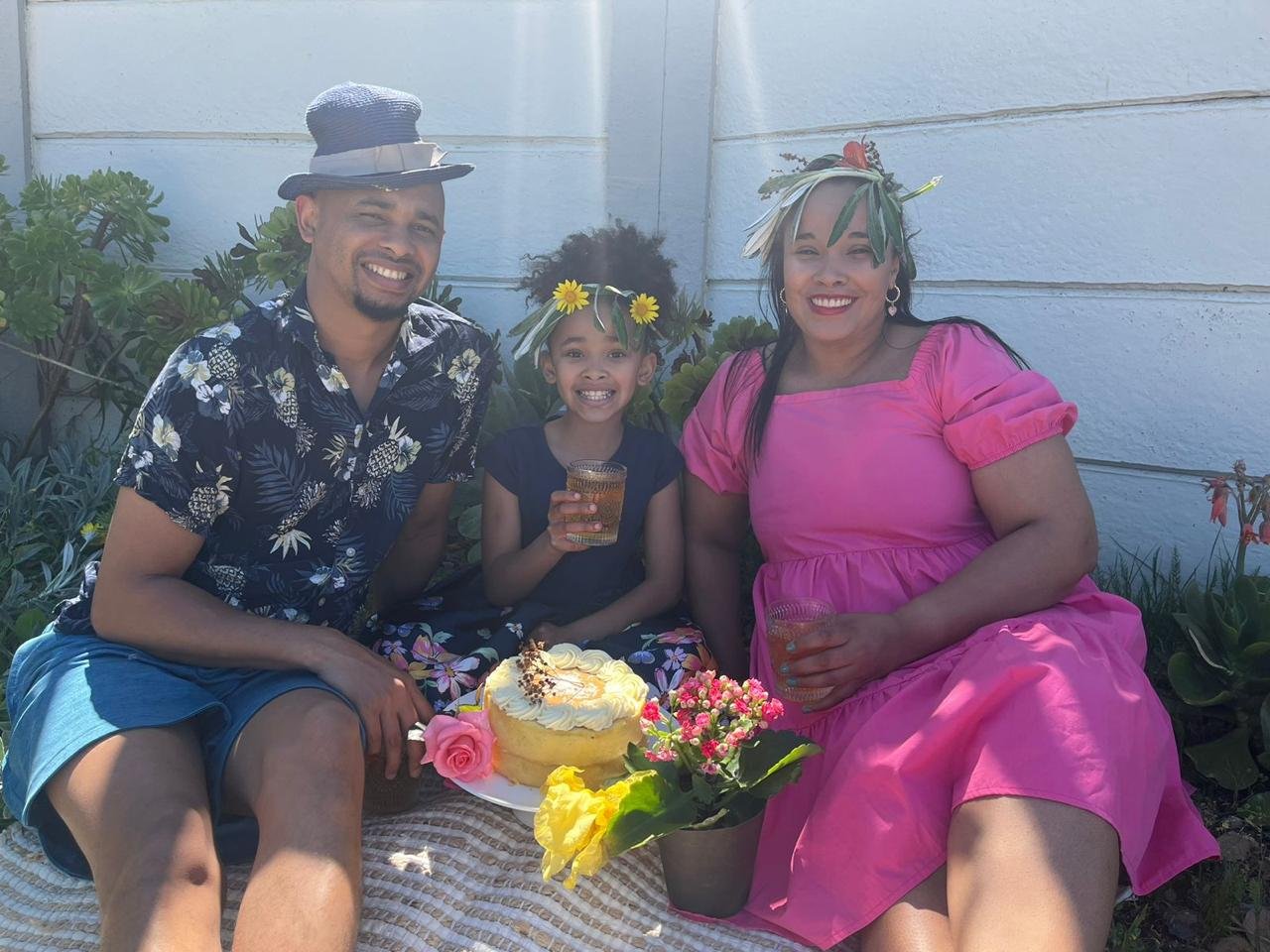 Family celebrating outdoors with a birthday cake, flowers, and drinks. All are smiling, wearing floral and tropical attire with flower crowns or hats, in front of a white wall with greenery.