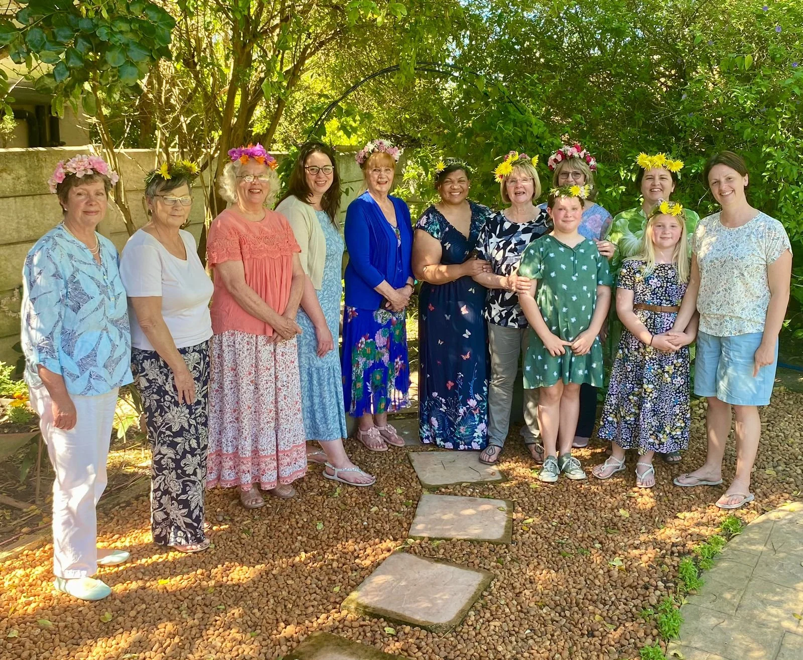 Group of women and girls outdoors, wearing flower crowns, standing on a gravel pathway in a garden with green trees and bushes in the background.