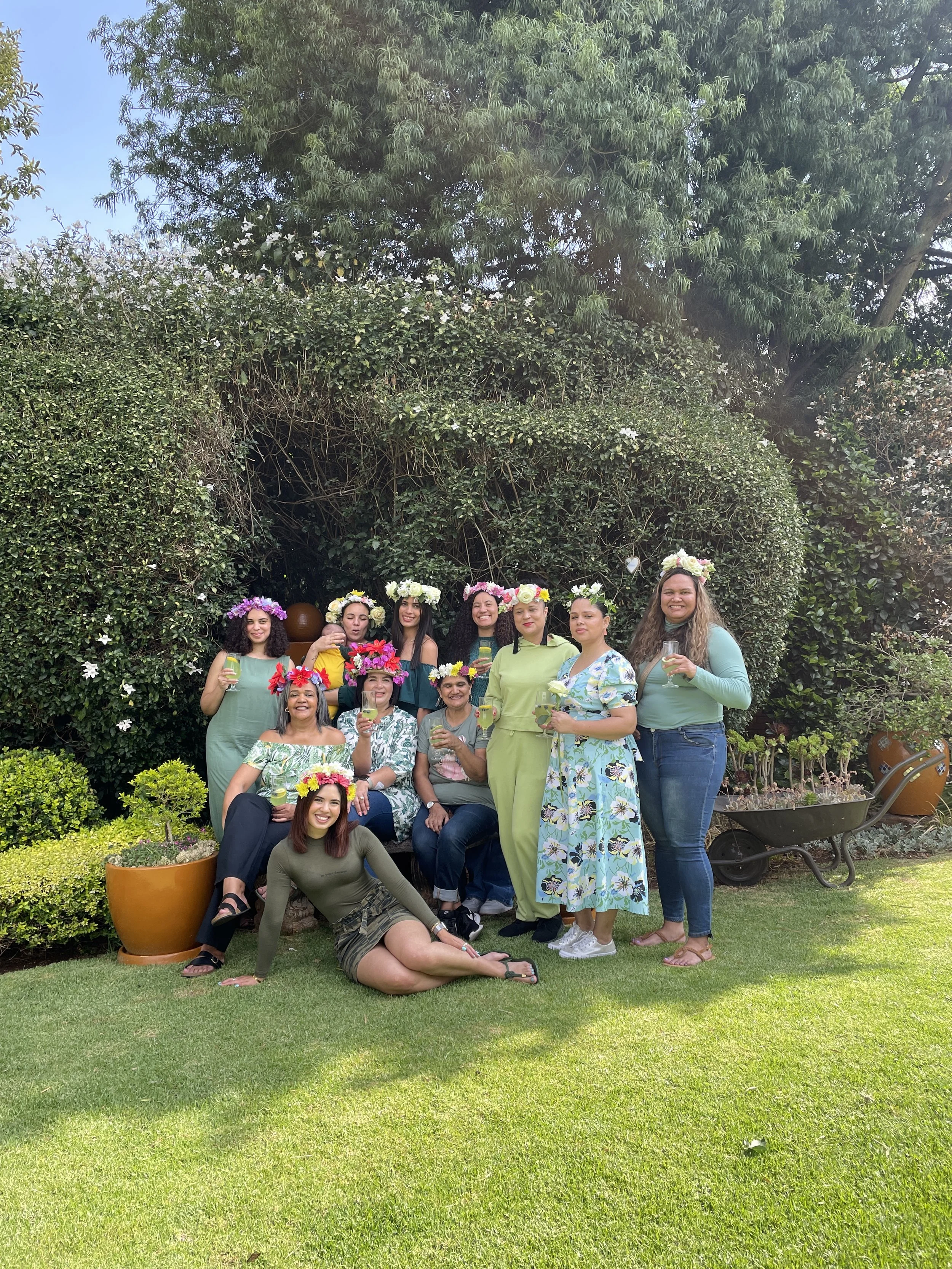 Group of women wearing flower crowns, holding drinks, posing in a garden with green bushes and trees in the background.