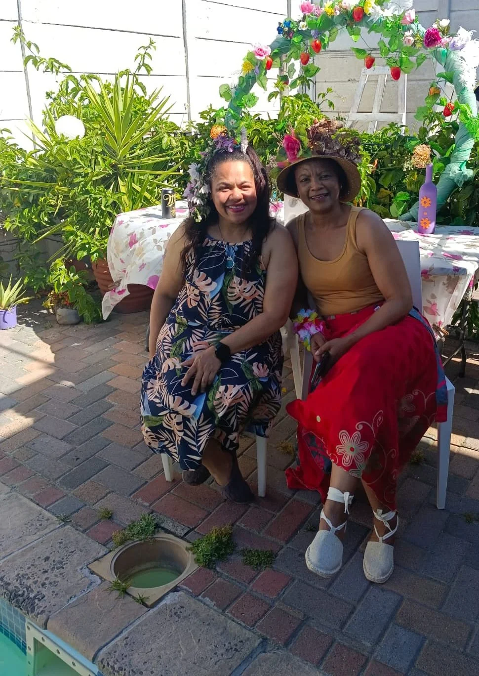 Two women sitting outdoors at a garden party, dressed in colorful summery clothing with flower accessories, smiling and enjoying the sunny day with a decorated table and greenery in the background.