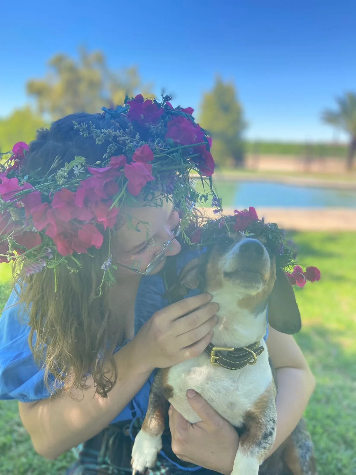 A woman with long hair and wearing glasses, decorated with a crown of flowers, is holding a happy brown and white puppy outdoors on a sunny day near a pond.