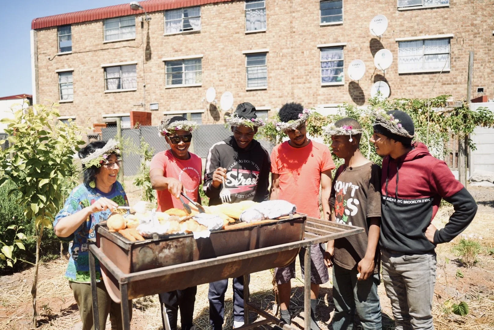 A group of six people, wearing flower crowns, stands together outdoors near a grill filled with food, smiling and chatting, with a brick apartment building in the background.
