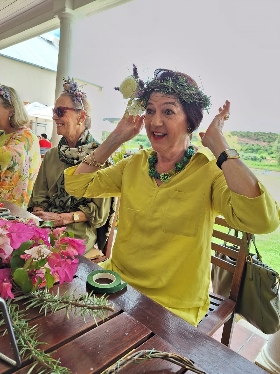 A woman wearing a yellow dress and a floral crown is adjusting or putting on a hat made of greenery and flowers, seated at a table with pink flowers and greenery, with other women nearby, in a bright outdoor setting.