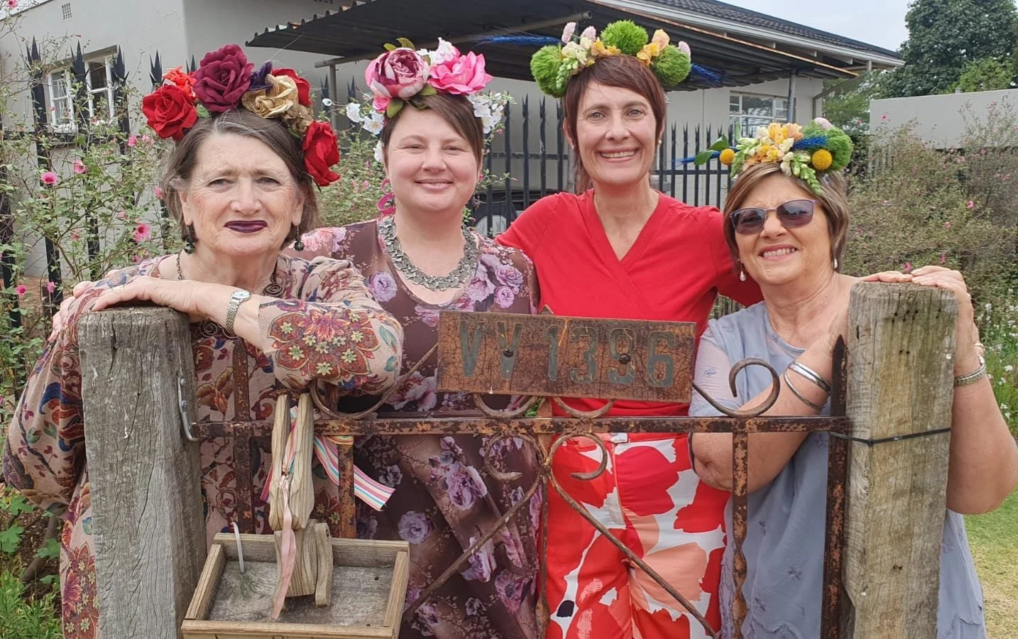Four women wearing floral crowns and colorful clothing, standing behind a wooden fence with a rusty metal sign that has the characters VY 1396. They are smiling and in a garden with flowers and bushes in the background.