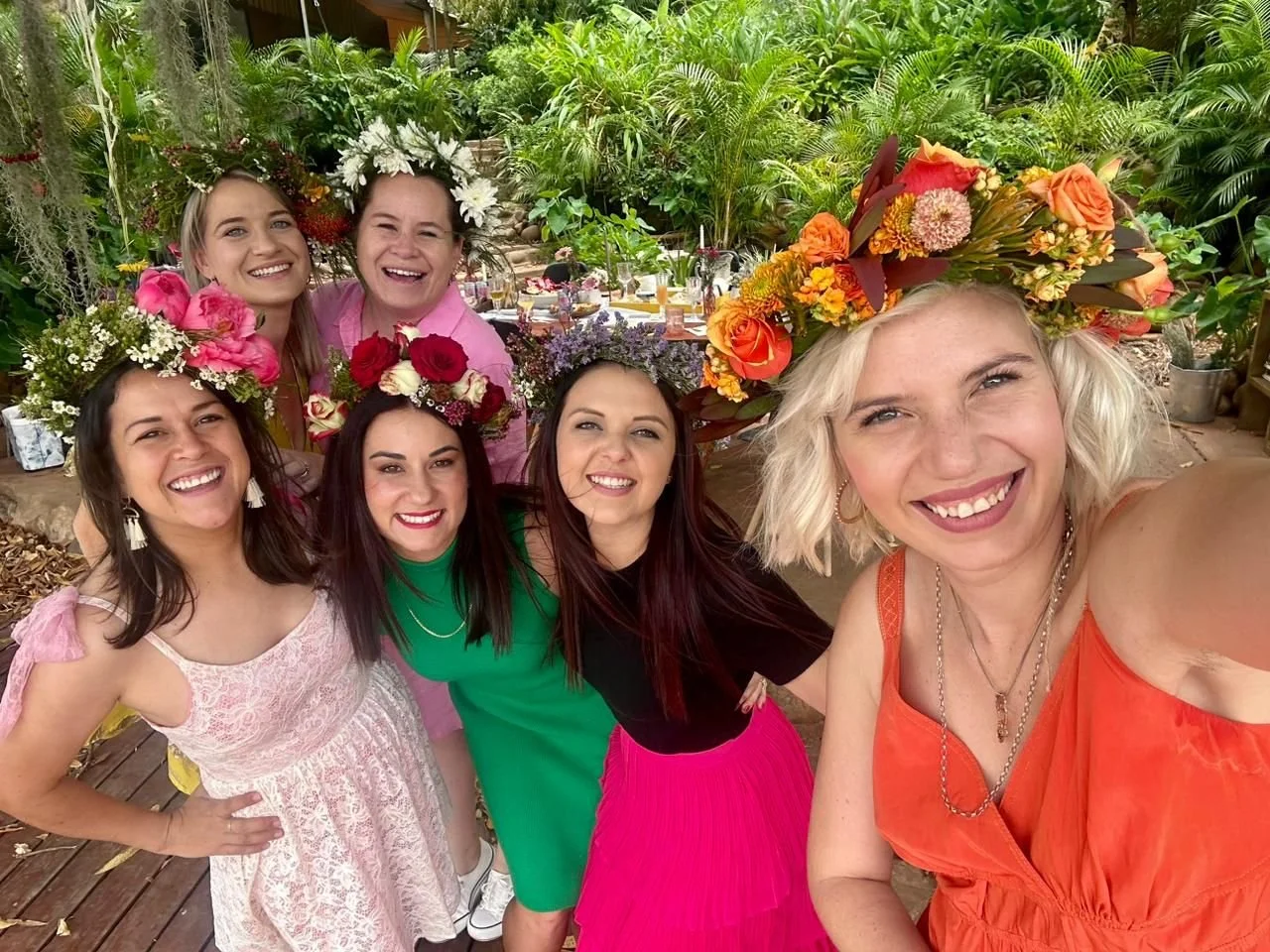Six women wearing floral crowns, taking a selfie at a garden party with lush greenery and decorated tables in the background.