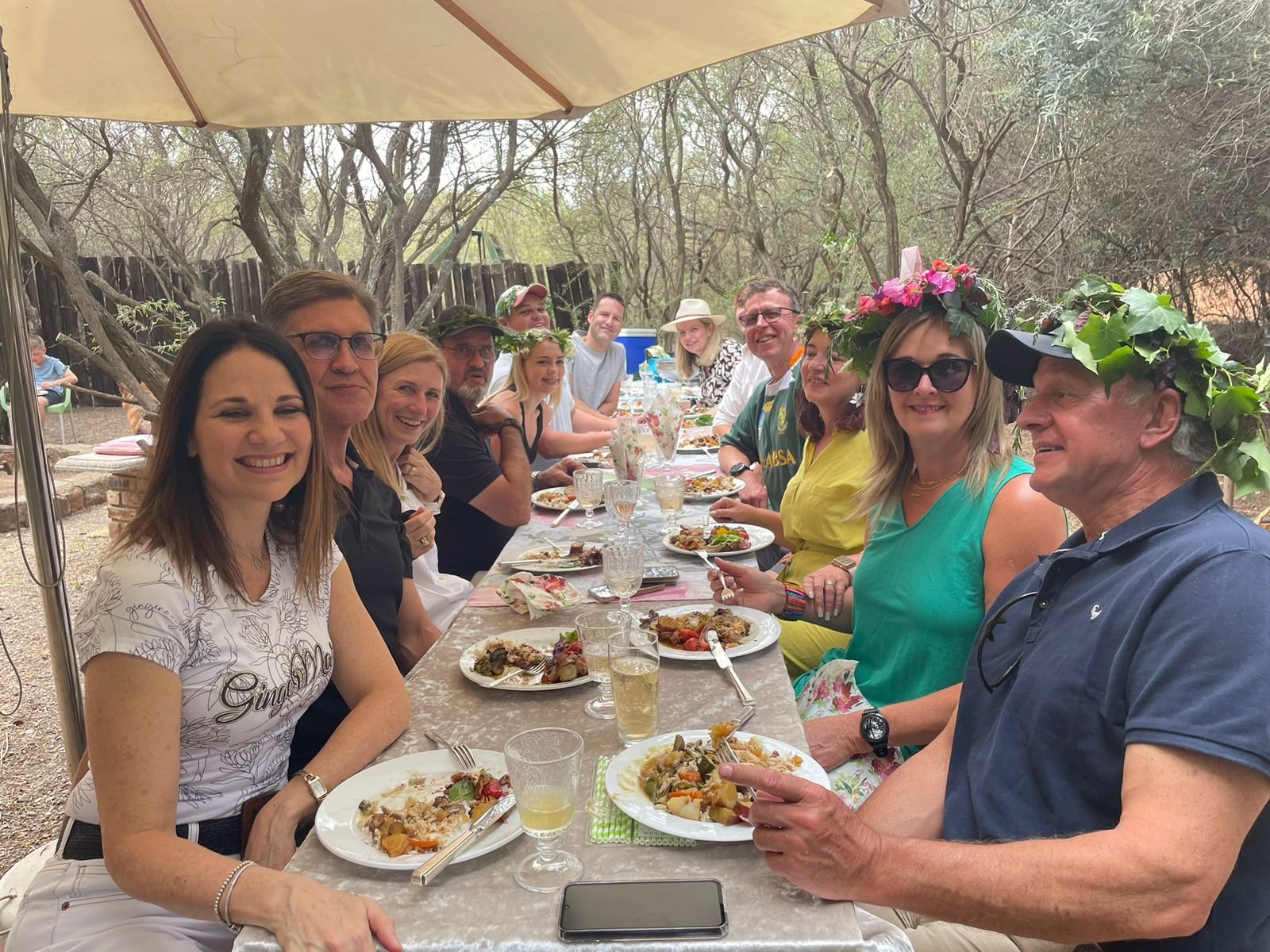 Group of people enjoying a meal outdoors at a long table, some wearing floral crowns and sunglasses, under a large umbrella in a wooded area.
