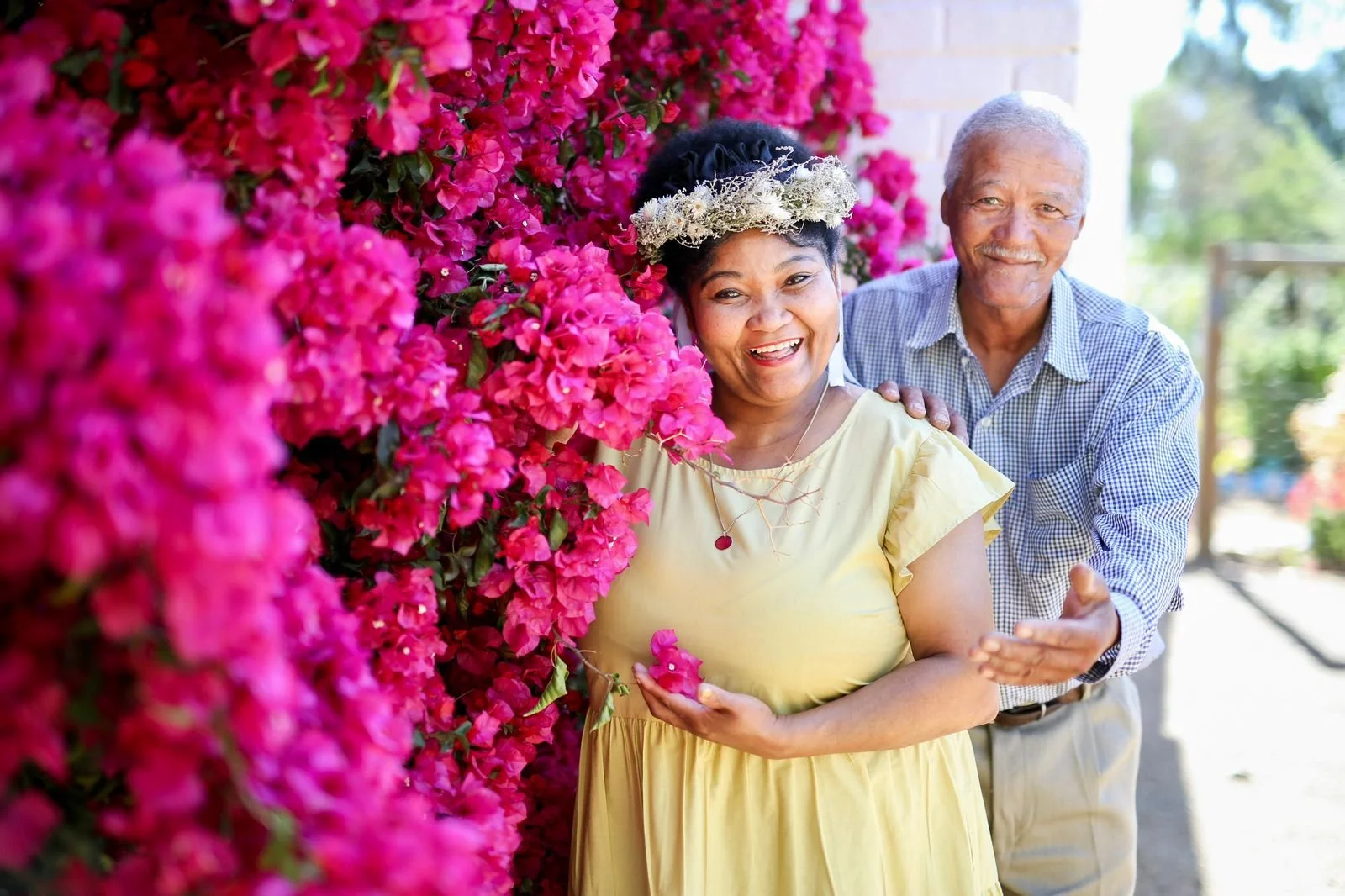 An elderly couple smiling and enjoying a moment in front of a vibrant pink bougainvillea flowers. The woman is wearing a yellow dress and floral crown, and the man stands behind her with a gentle smile, holding her shoulder.