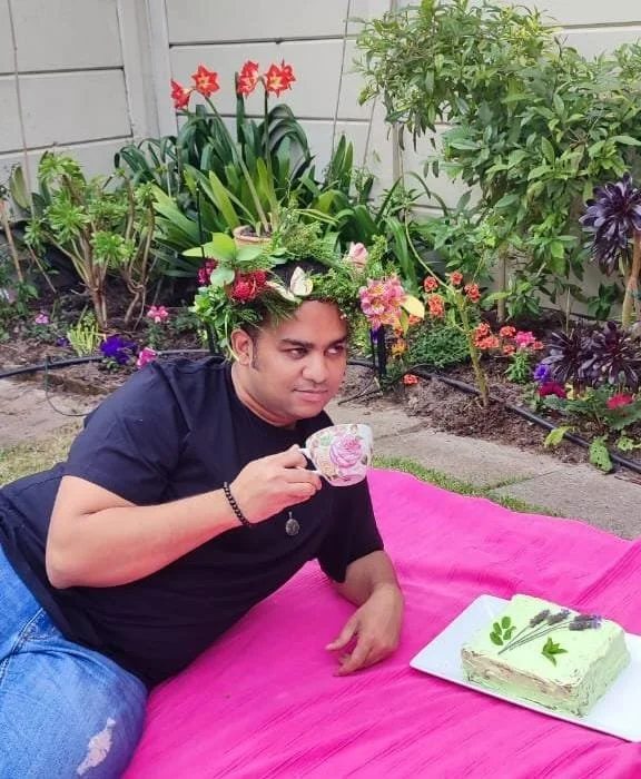 A person sitting on a pink blanket in a garden, wearing a flower crown and holding a teacup, with a decorated cake on a plate nearby.