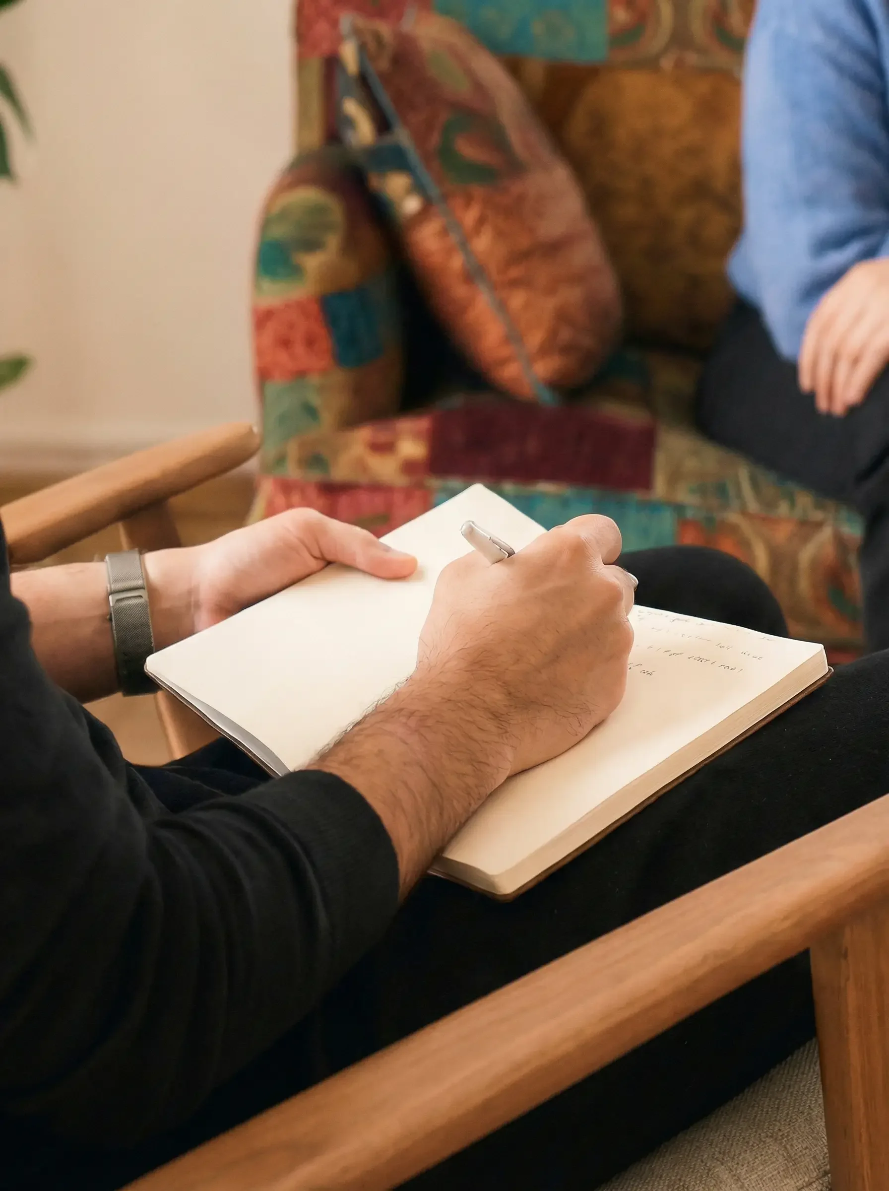 Nahaufnahme einer Person, die auf einem Holzstuhl sitzt und in einem Notizbuch schreibt, während eine andere Person im Hintergrund sitzt.