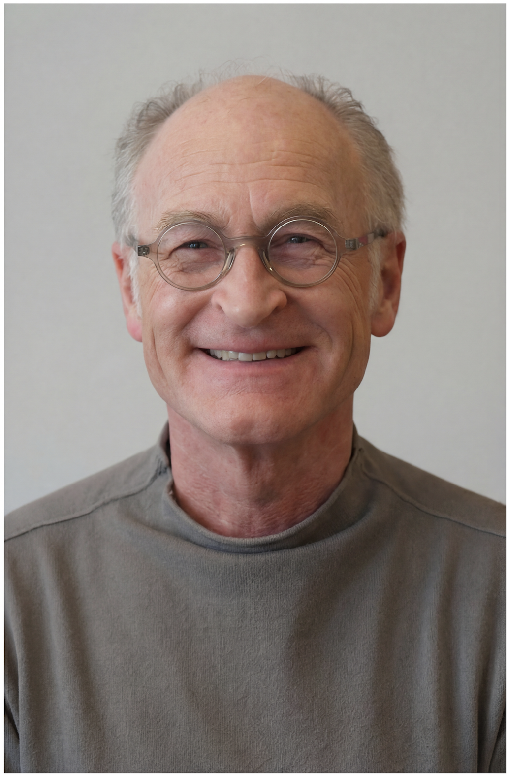 Ein älterer Mann mit Brille, lächelnd, trägt einen grauen Pullover, steht vor einem neutralen Hintergrund.