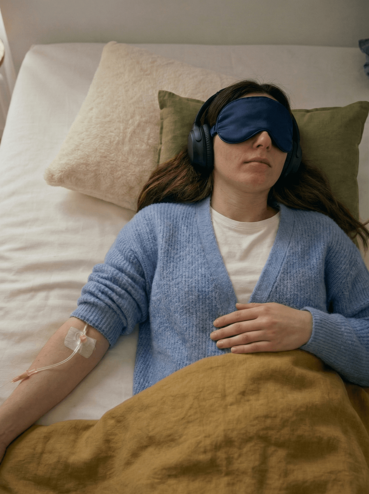 Frau liegt auf einem Bett, trägt eine Schlafmaske, Kopfhörer und eine blauen Pullover. Sie hat einen Zugang für eine Infusion im Arm.
