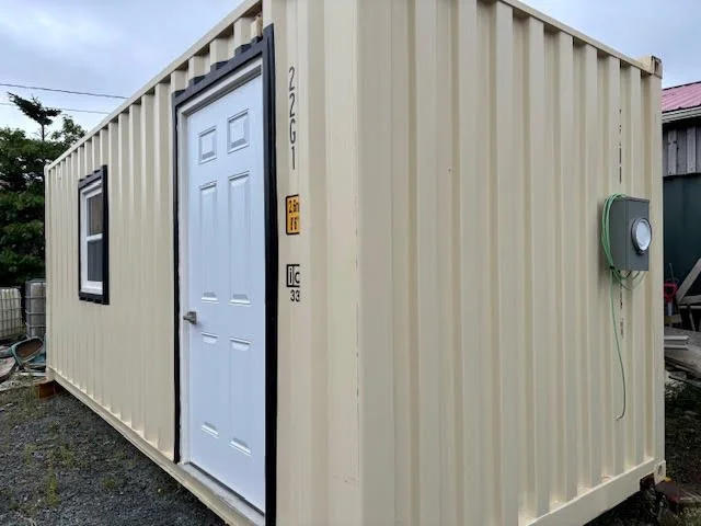 A beige shipping container converted into a small building with a white door, a window, and an electrical meter on the exterior.
