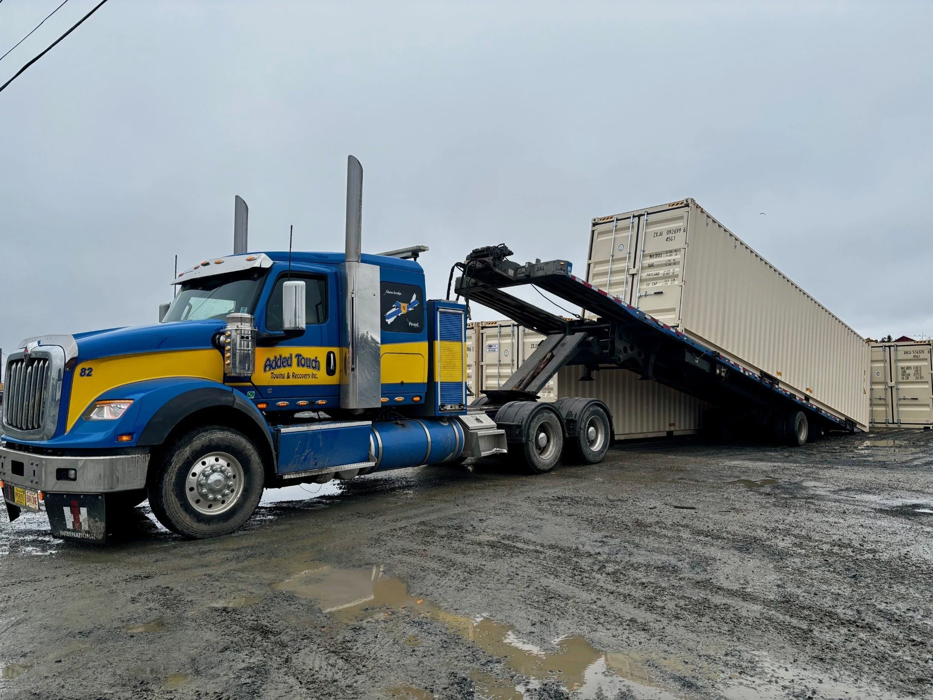 A blue and yellow tow truck with 'Added Towed' on the side, lifting a shipping container on a muddy, wet ground under a cloudy sky.