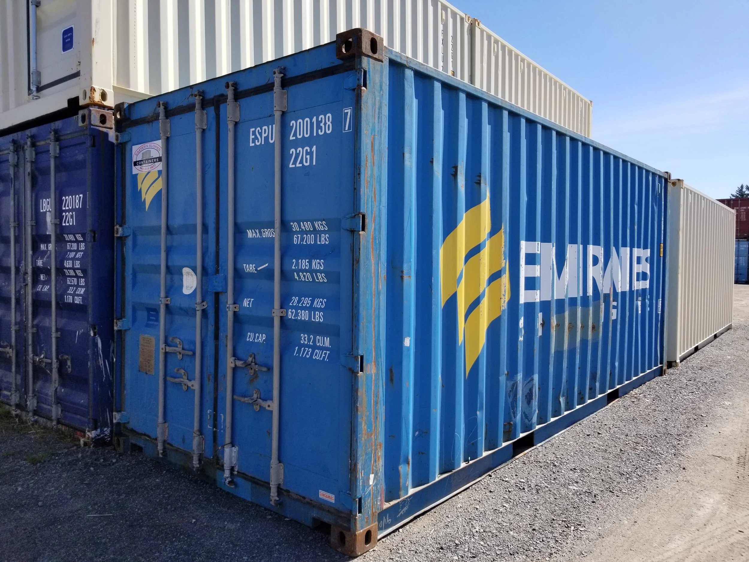 A blue shipping container with the word "EMIRATES" and a logo, placed on gravel outdoors with other containers in the background and a clear sky above.