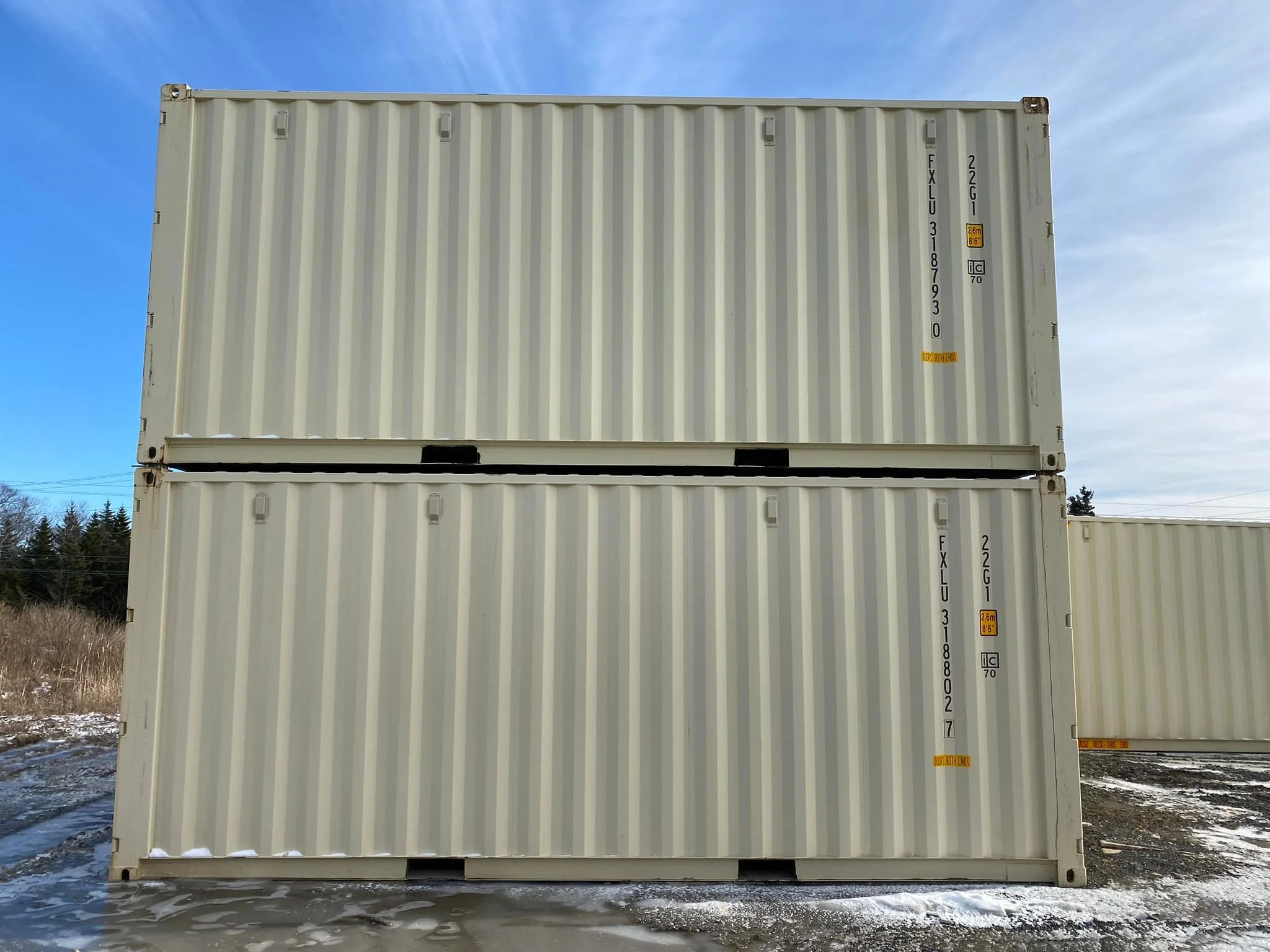 Two stacked beige shipping containers outdoors under a cloudy sky with a snowy ground and trees in the background.