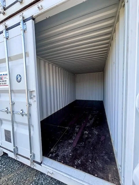 Empty shipping container with white corrugated metal walls and slanted roof, black flooring, and some debris inside.