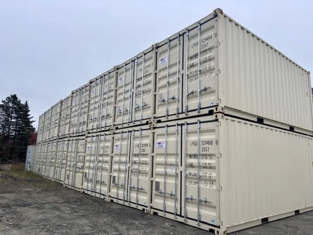 Two stacked beige shipping containers outdoors on a gravel surface with trees in the background.