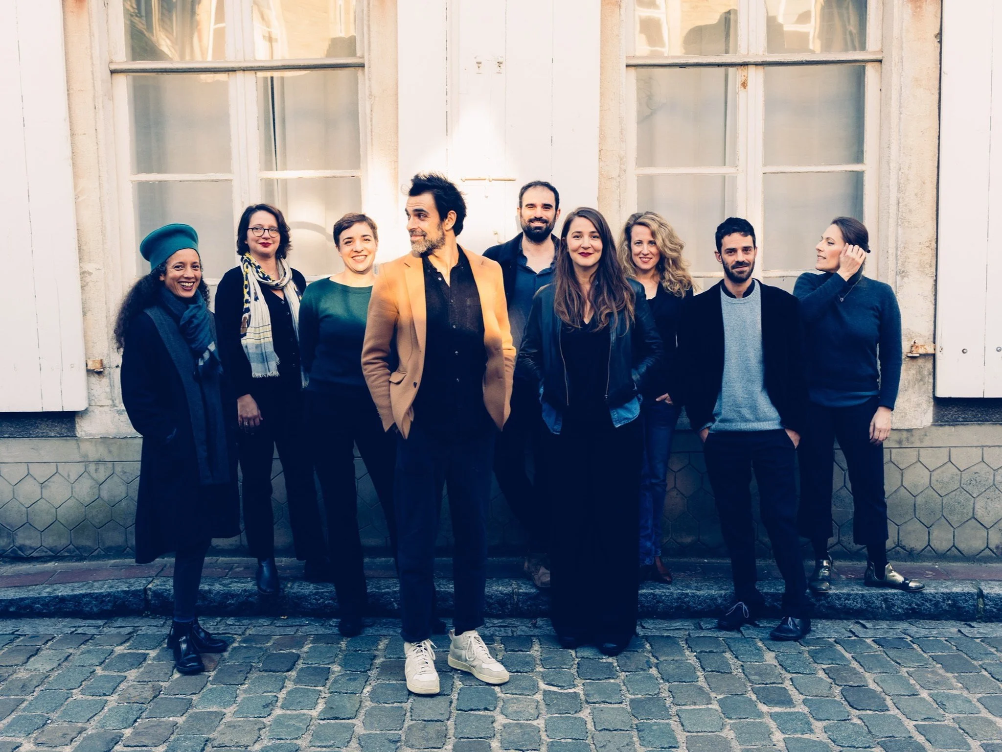 Groupe de personnes posant ensemble devant un bâtiment avec des fenêtres blanches, certaines souriantes, sur un sol pavé.
