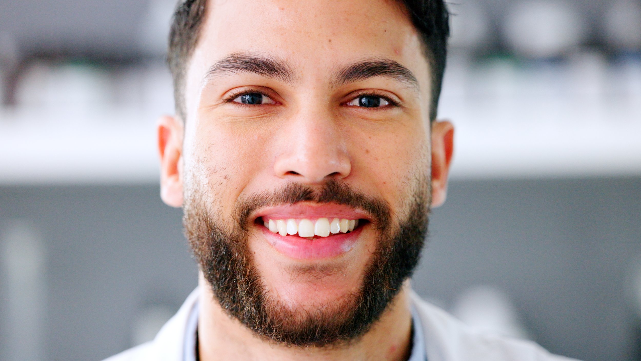 Close-up of a smiling man with a beard and short dark hair, wearing a white coat in a bright indoor setting.