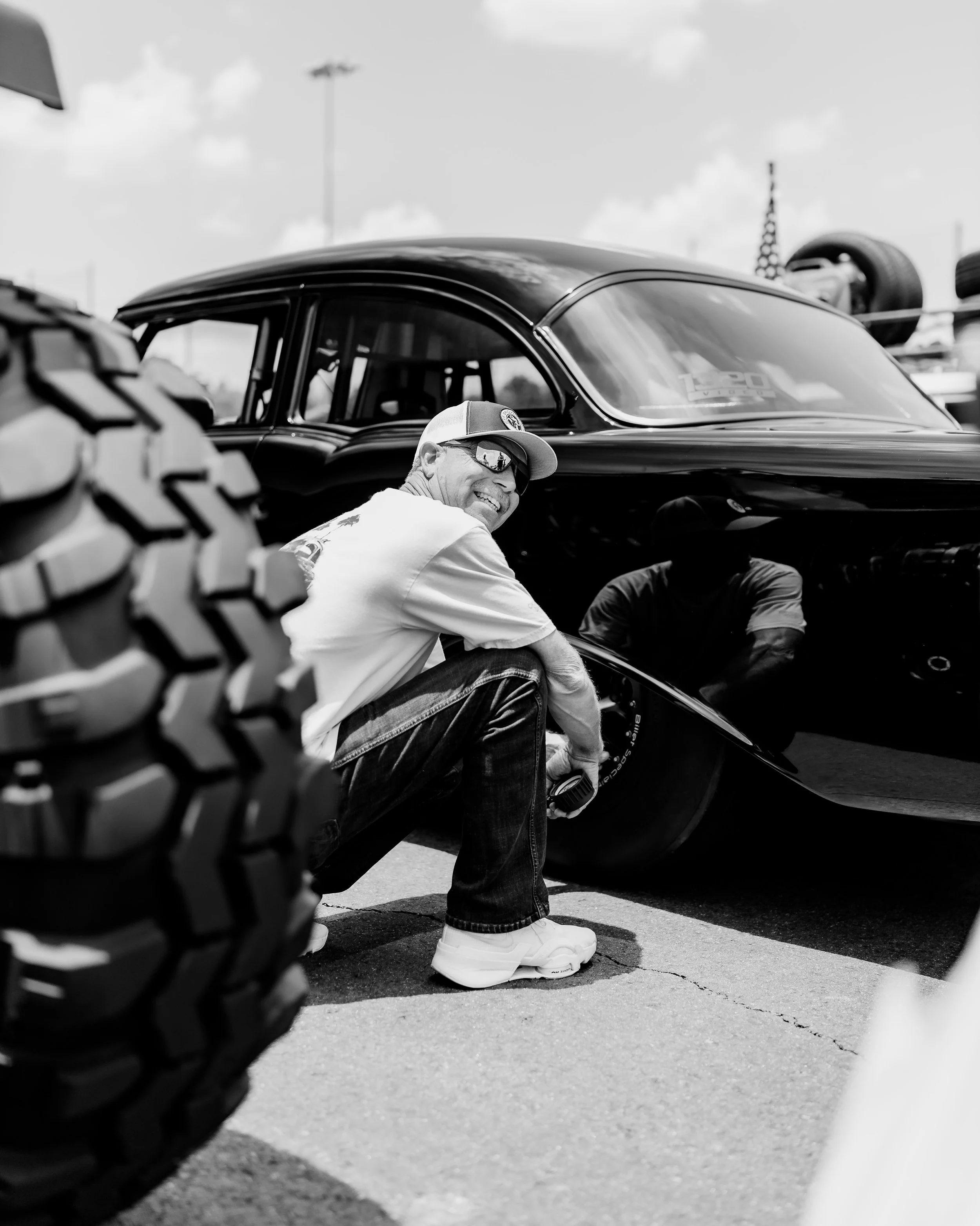 A man wearing sunglasses and a cap squats and smiles next to a vintage race car, with a blurred tire in the foreground.