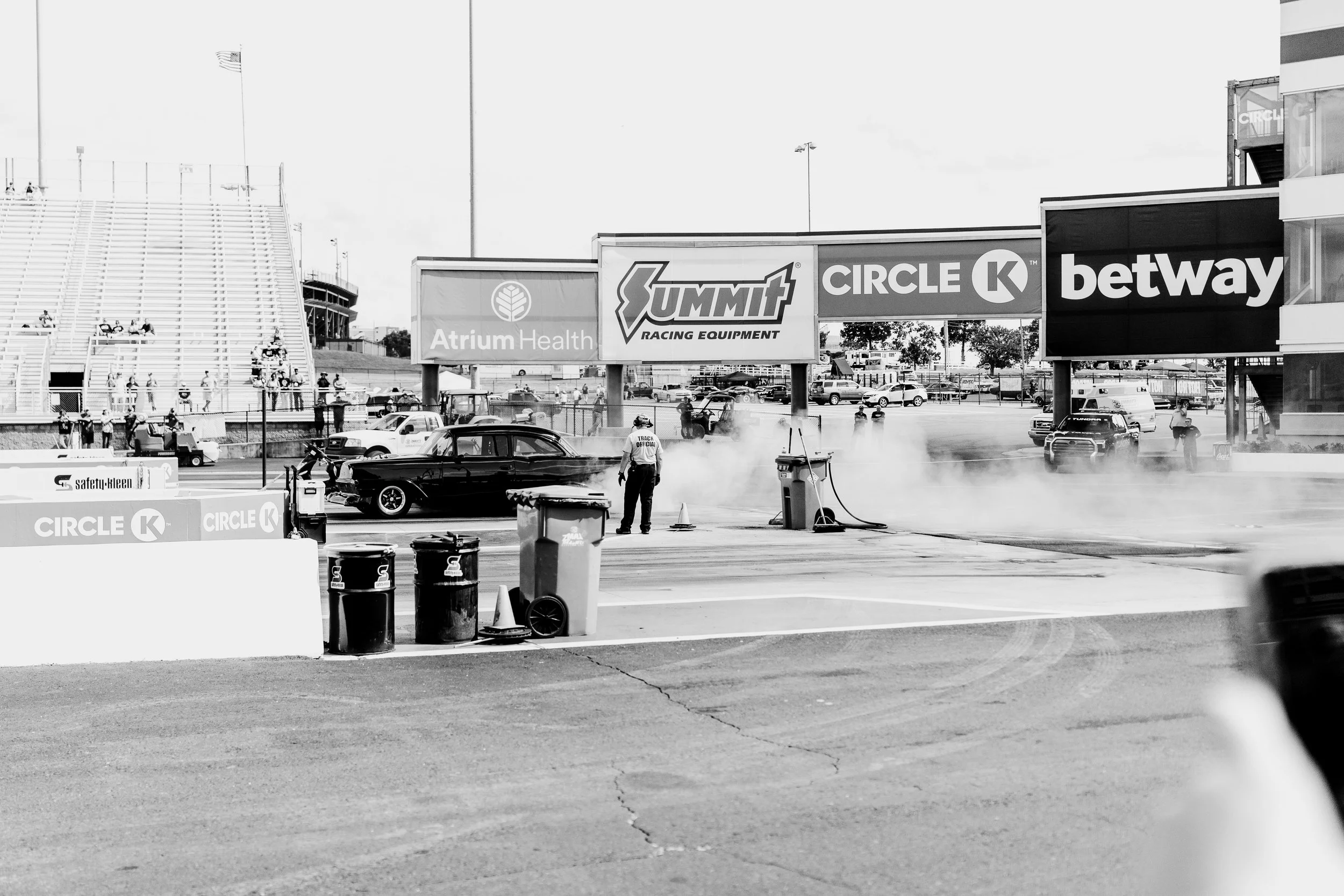 A drag racing event at a racetrack with a black classic car starting to burn rubber as smoke billows from the tires. There are people observing and officials working near the track, with advertising banners and empty grandstands in the background.