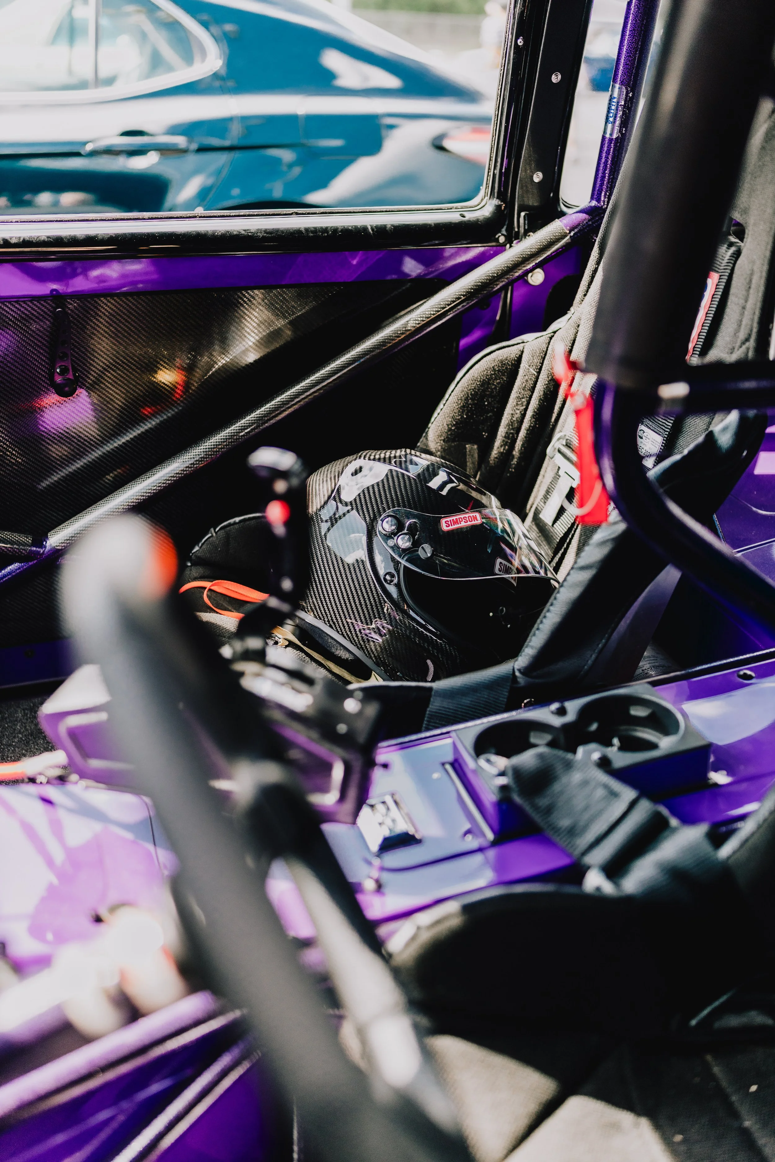 View of the interior of a race car showing a black helmet with red and white Simpson logos, a racing seat with harness, and part of the purple exterior and window.