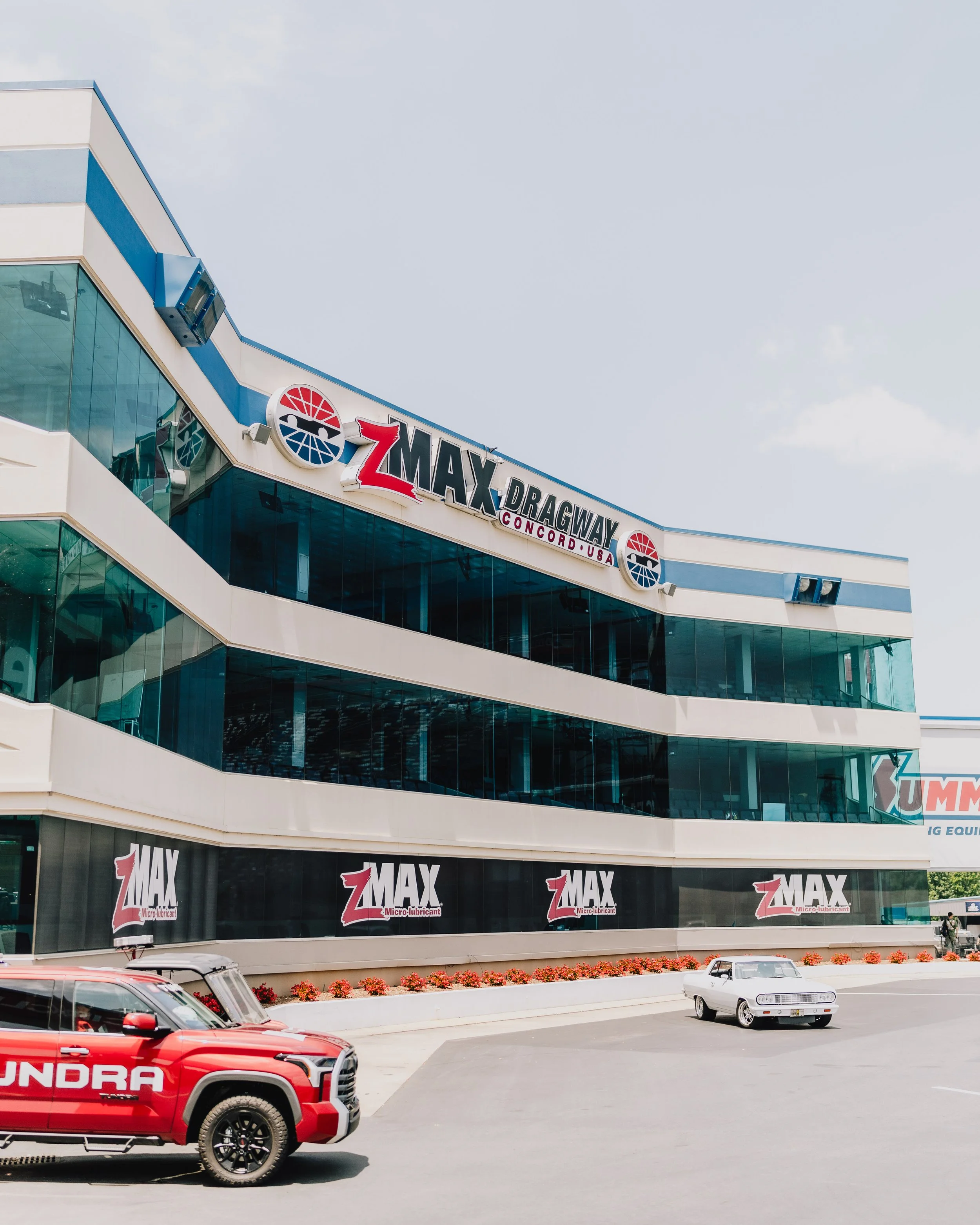 Exterior view of ZMAX Dragway at Concord, USA, showing the multi-story building with glass windows, signage, and parked trucks in the foreground.
