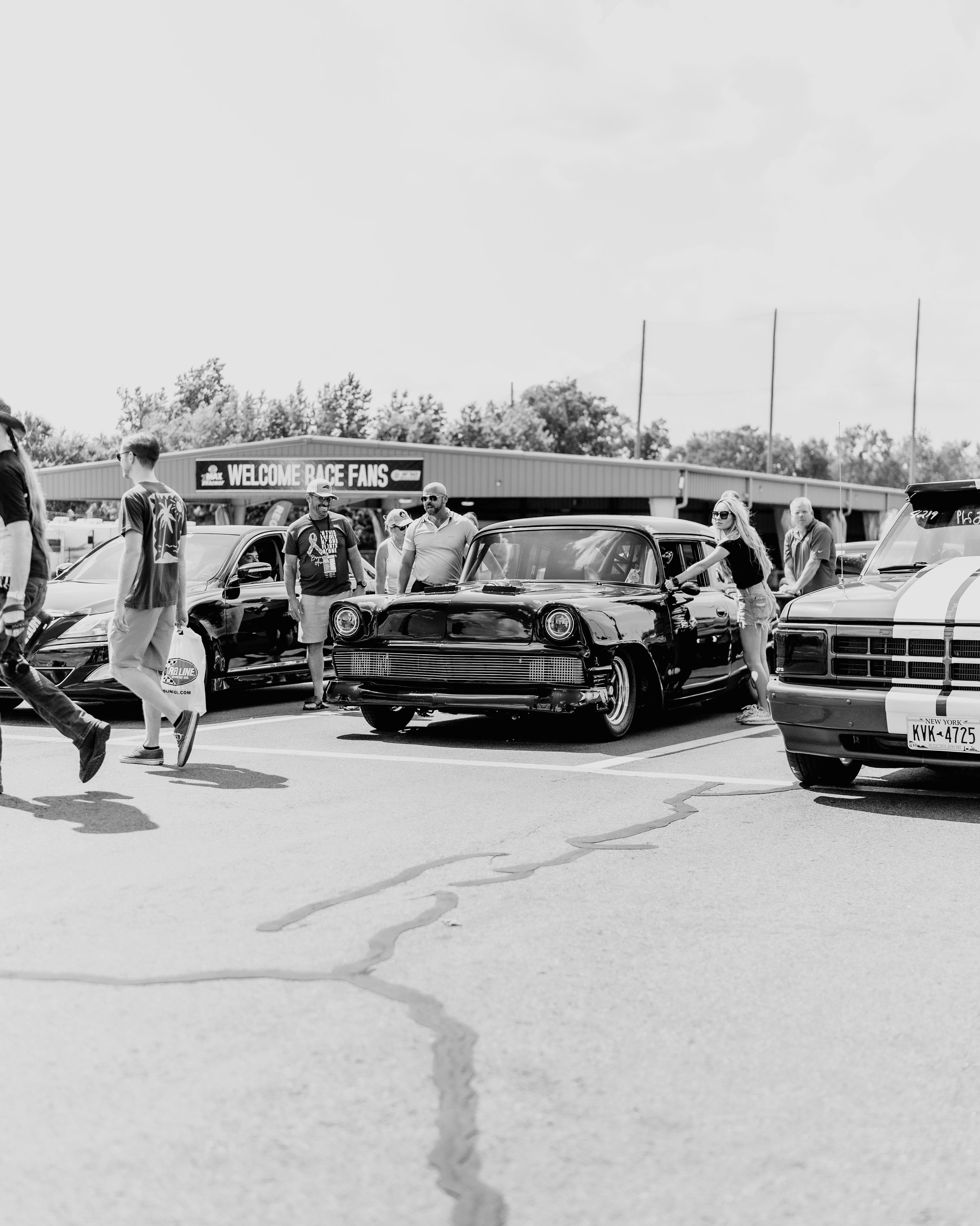 Fans gather around cars at a racing event, with a sign in the background saying "WELCOME RACE FANS".