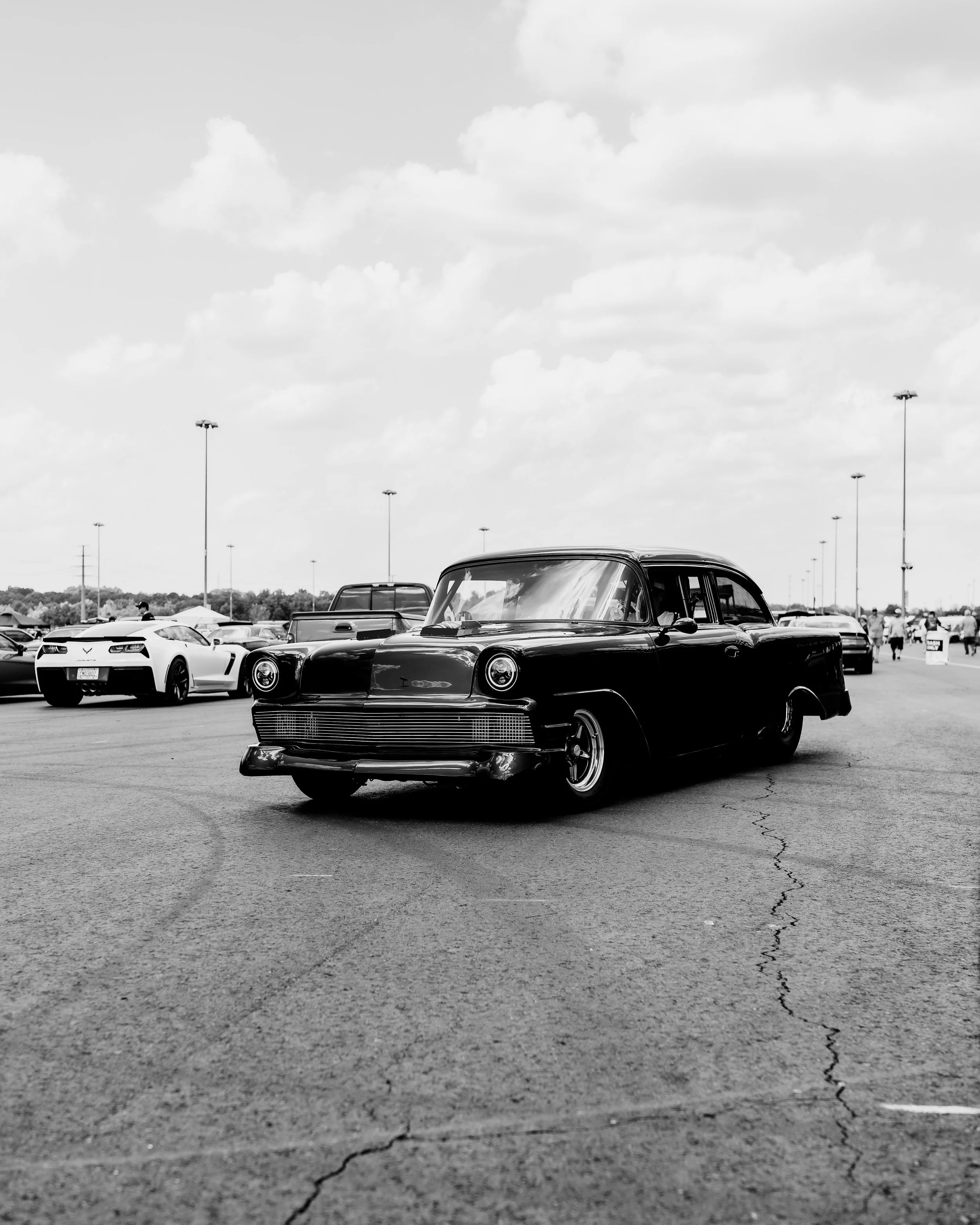 Black vintage car parked on asphalt at car show with modern cars and people in the background, under cloudy sky.