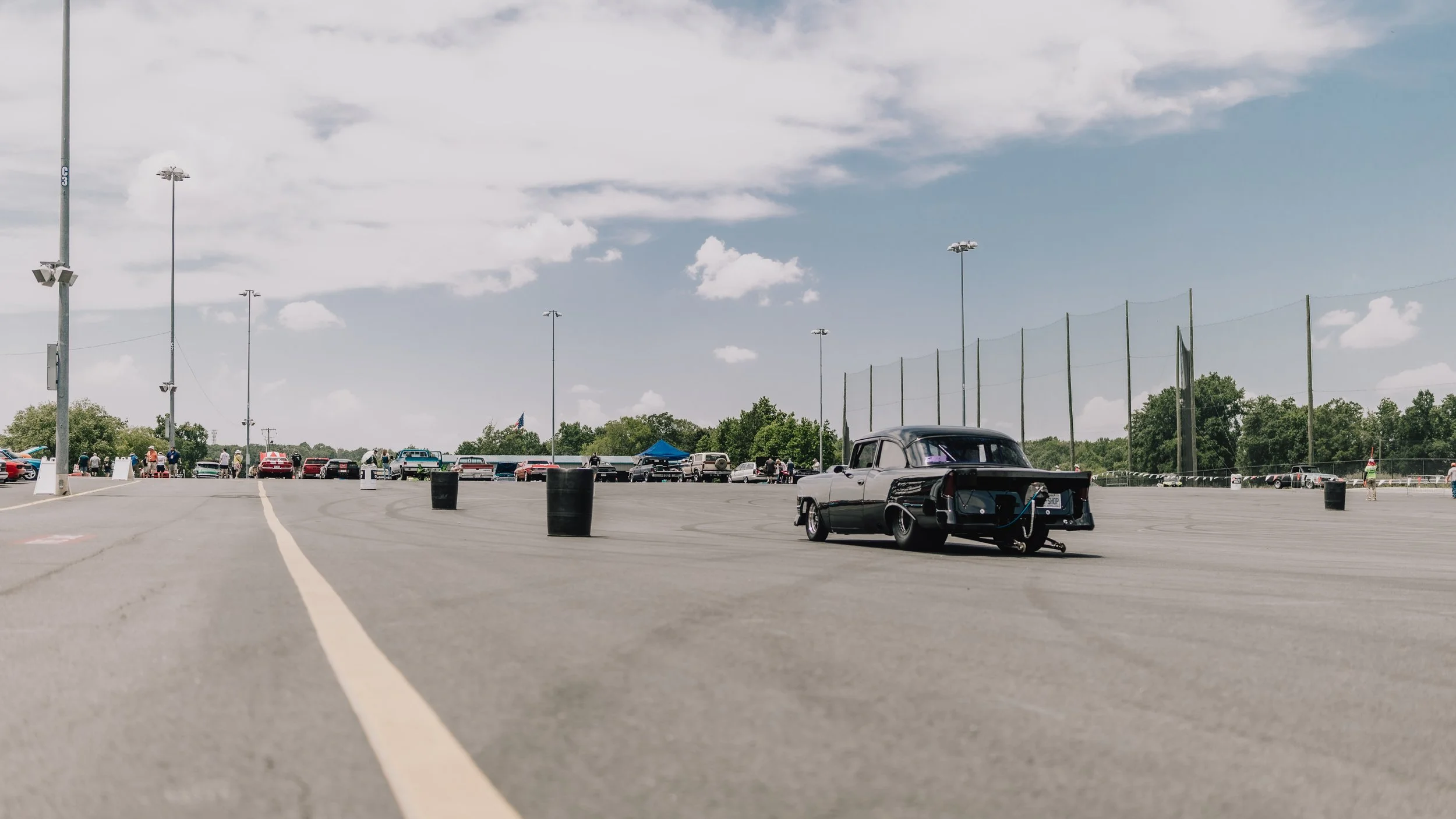 Vintage black car in a large outdoor parking lot with other vehicles in the background, under a partly cloudy sky.