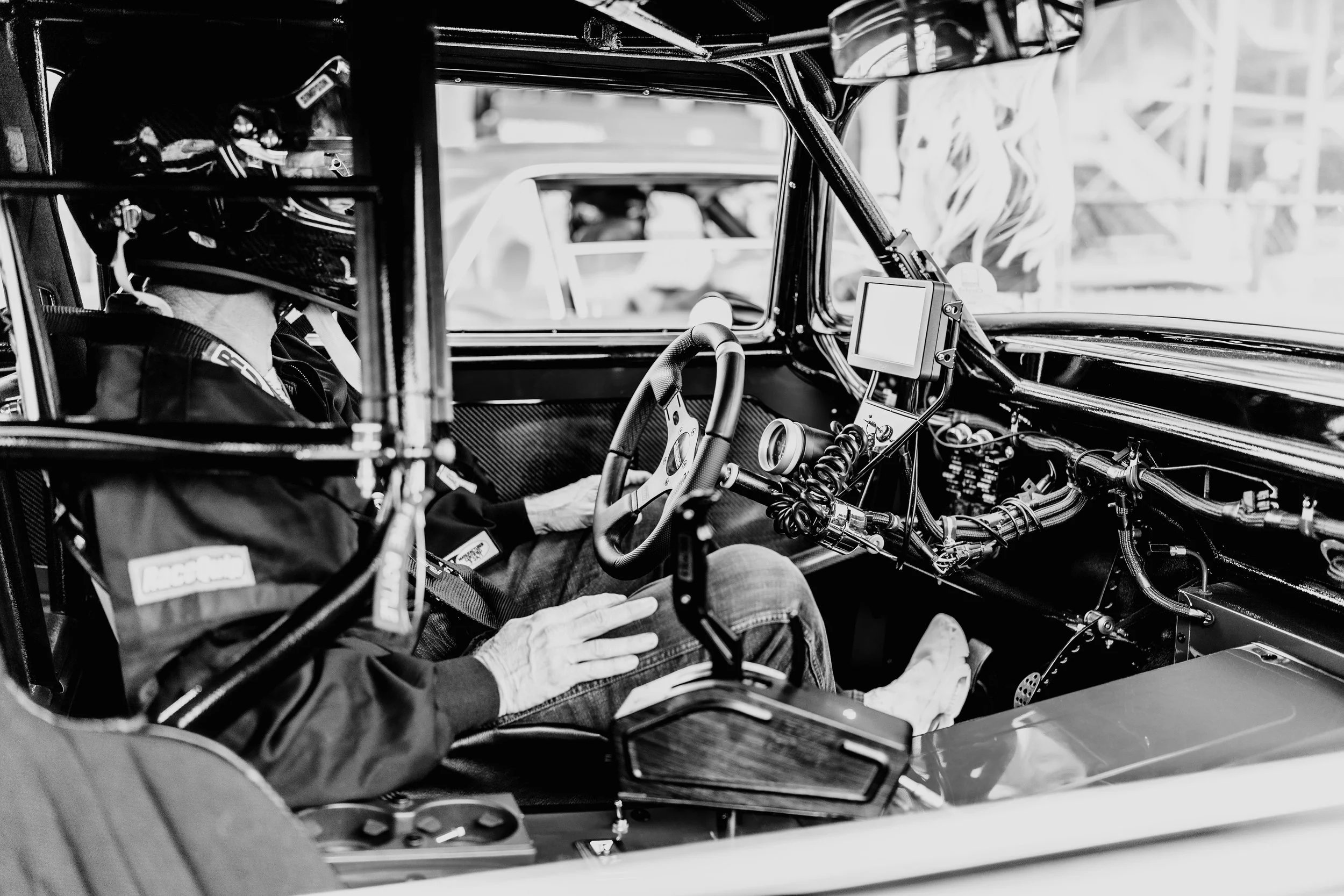 A race car driver sitting in the cockpit of a race car, wearing a helmet and racing gear, with various equipment and controls visible inside the car.