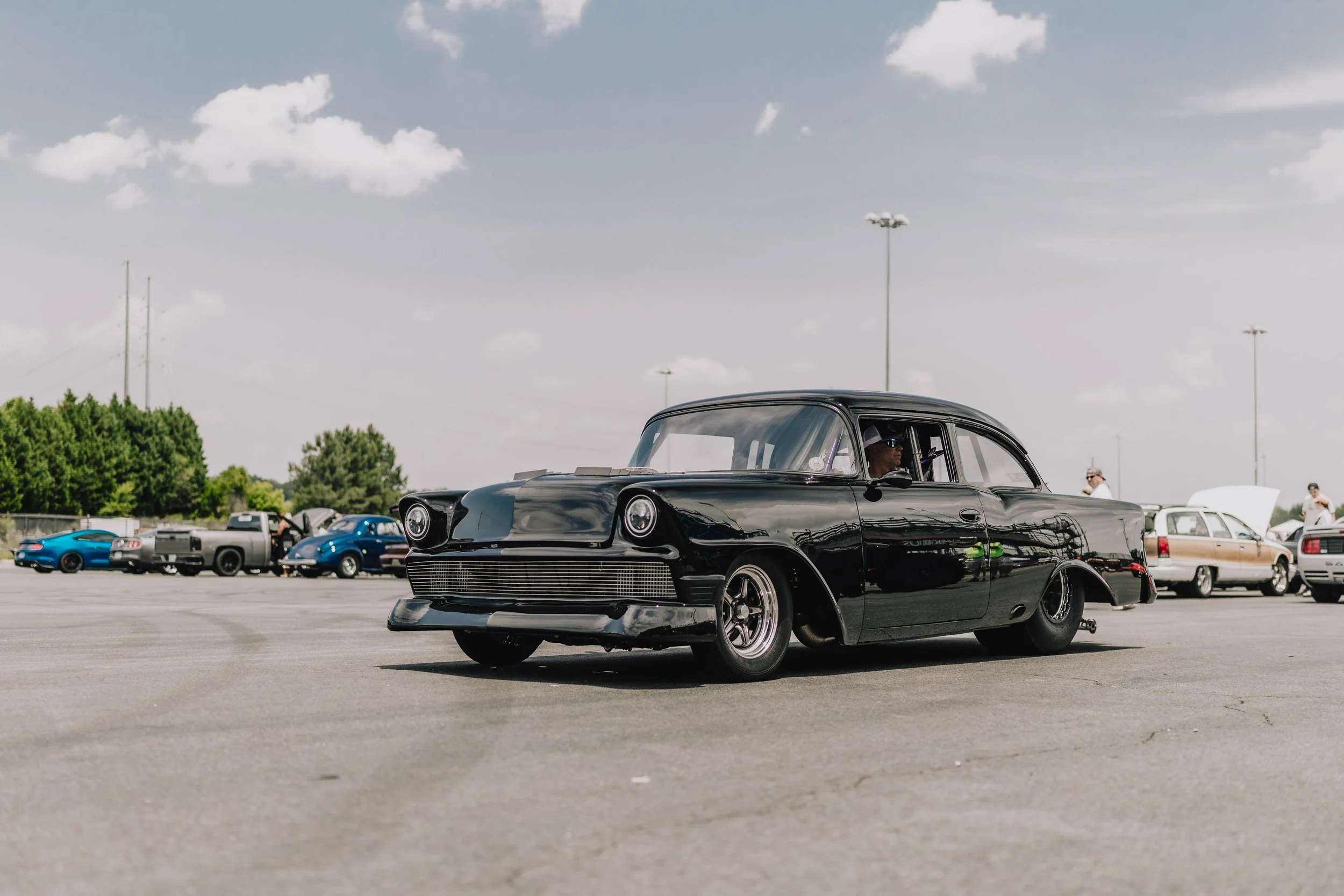 A black classic vintage car on display at a car show, with other classic and modern cars in the background on a sunny day with a cloudy sky.