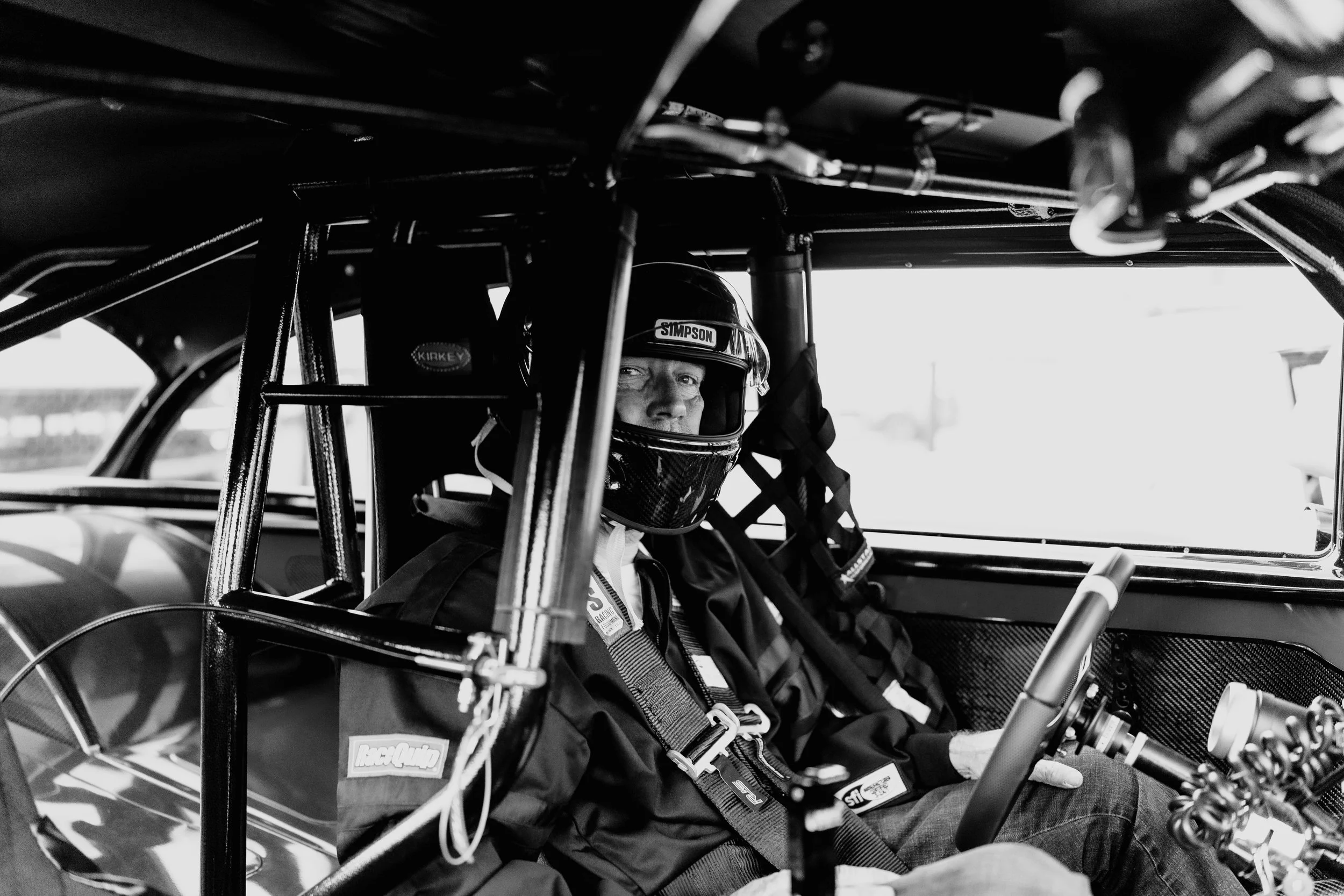Black and white photo of a race car driver sitting inside a race car, wearing a helmet and racing suit, looking towards the camera.