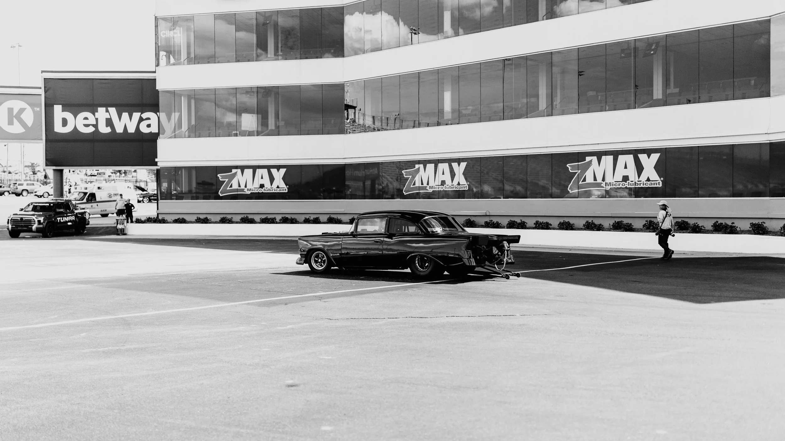 Black vintage car on a tow truck in a parking lot of a commercial building with Betway and Z-Max signs, people walking, and parked cars in the background.