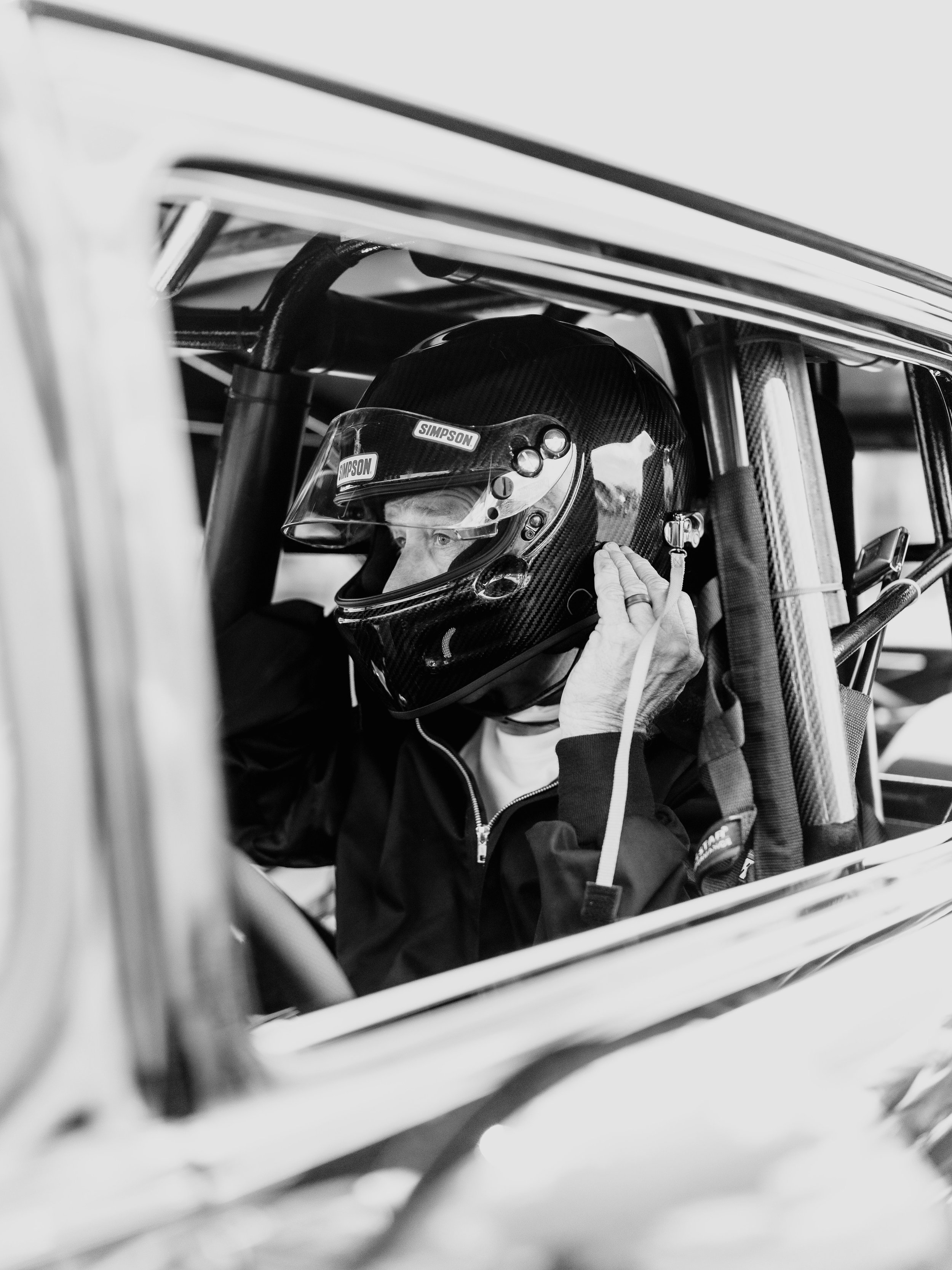 A race car driver wearing a helmet and suit, sitting inside a racing car, adjusting the helmet strap.