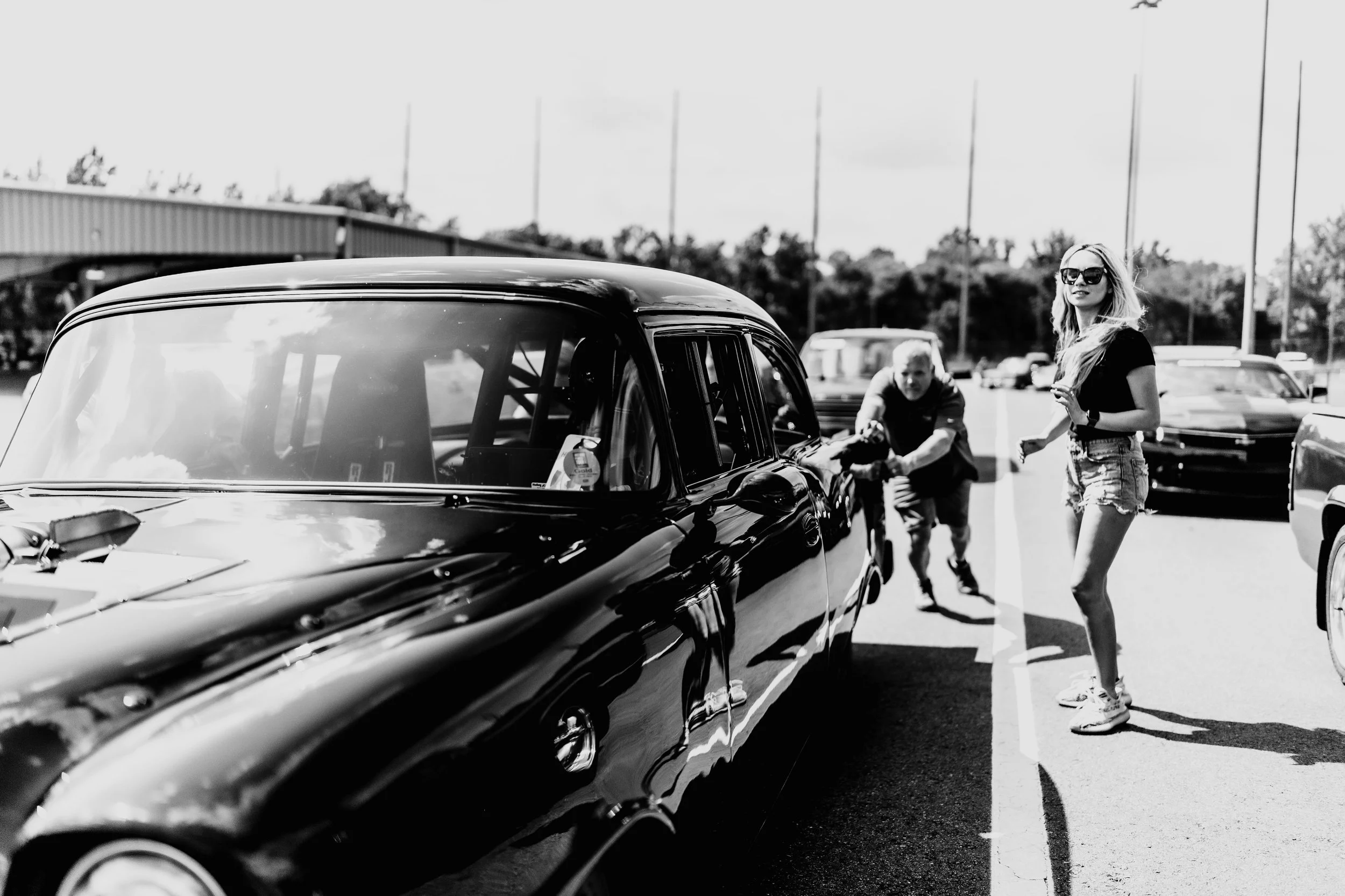 A woman with long hair, sunglasses, a black t-shirt, and denim shorts standing in a parking lot, with a young person pushing a bicycle behind her, beside a shiny black vintage car, and other cars in the background.