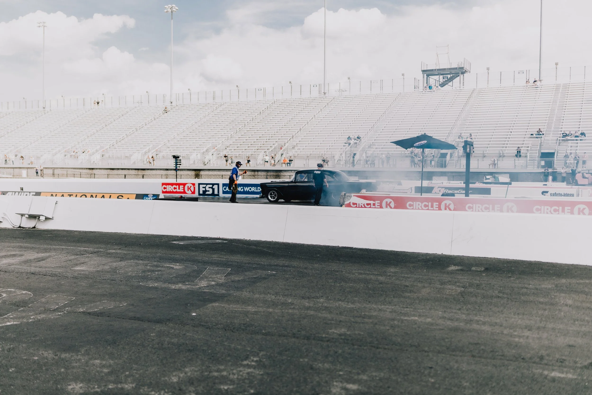A drag racing event with a classic black car on the track, two officials standing nearby, tire smoke in the air, and empty grandstands in the background under cloudy skies.