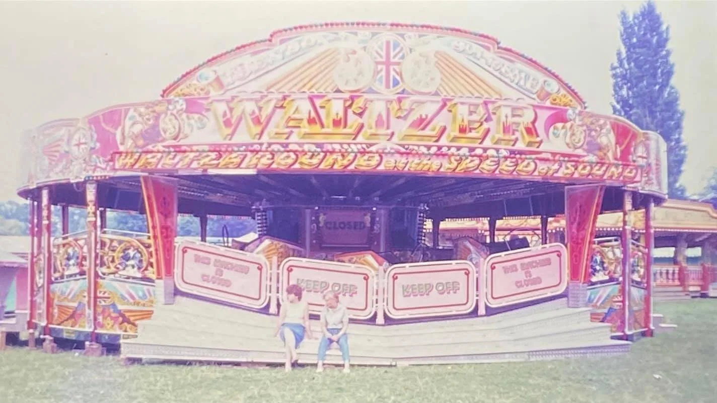 Vintage amusement park ride with colorful "Waltzer" signage, closed for maintenance, and two people sitting on the front ledge.