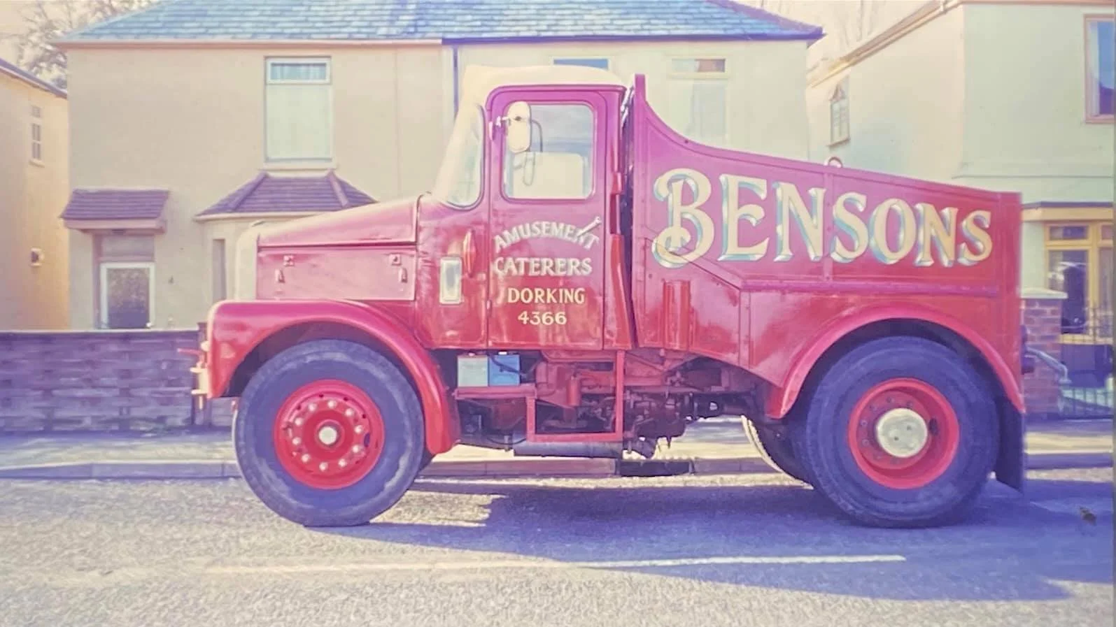 A vintage red truck parked on a street in front of a house, with the words "Bensons" and "Amusement Caterers Dorking 4366" painted on the side.