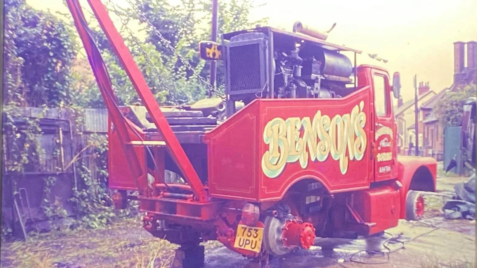 Red vintage tow truck with "Benson's" written on the side, missing rear wheels, in a yard with overgrown vegetation.