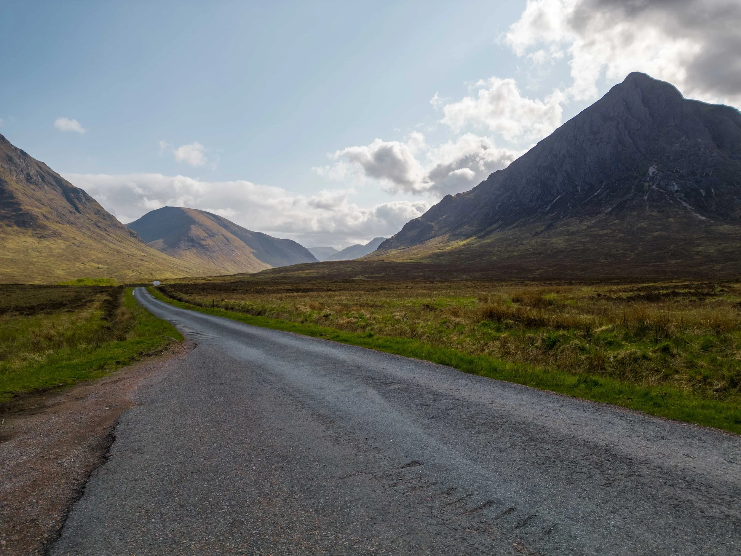 A winding road running through a green valley surrounded by tall mountains under a partly cloudy sky.