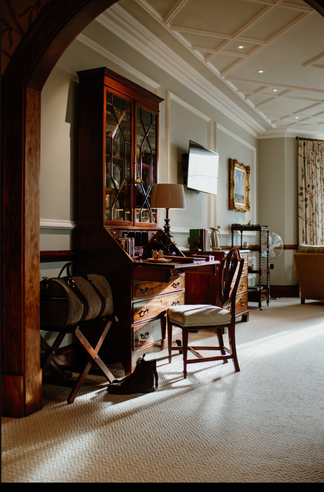 A cozy hotel or living room with a wooden desk, chair, and armoire, decorated with books, a lamp, and personal items, illuminated by natural sunlight.