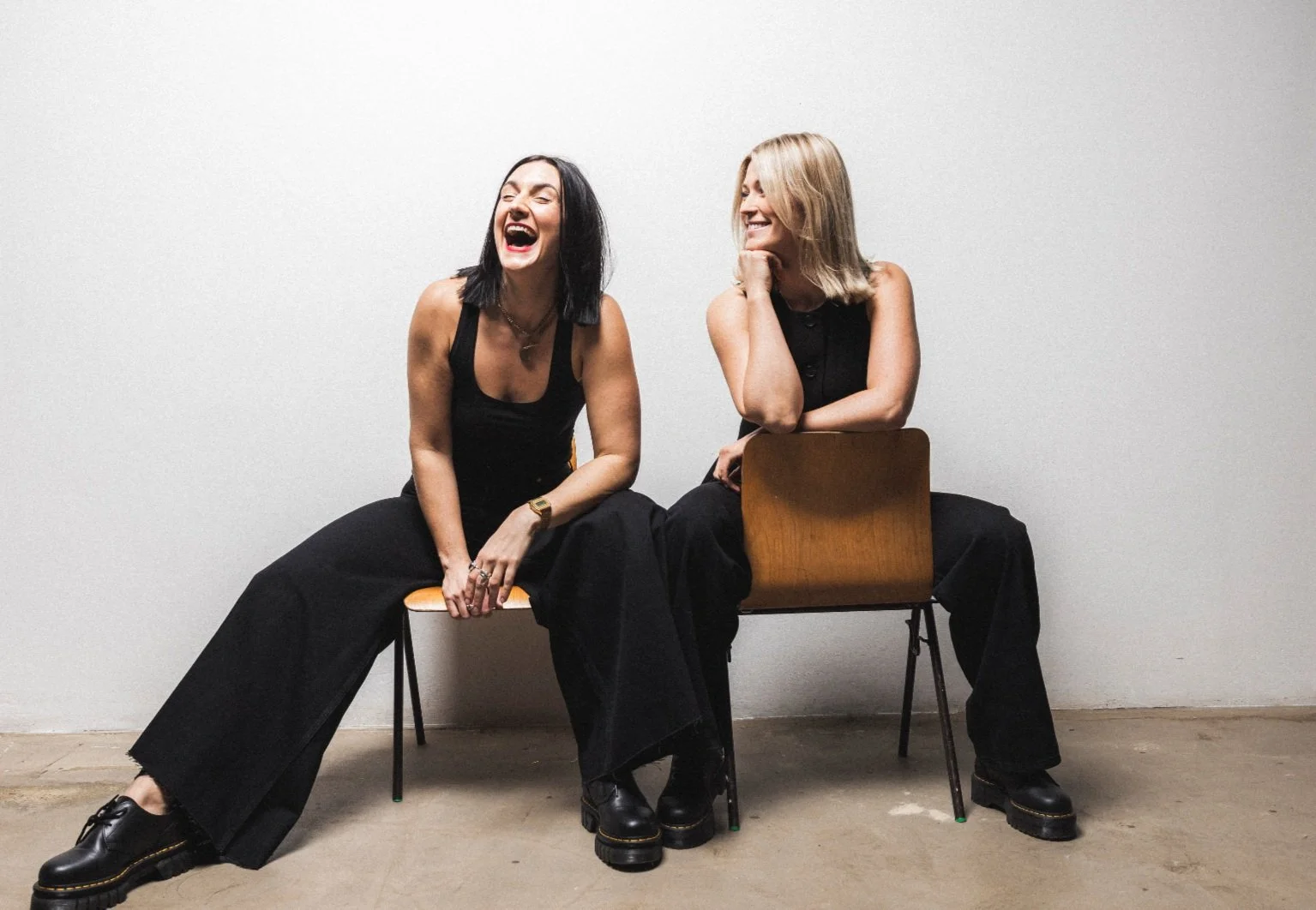 Two women dressed in black, sitting on chairs against a plain wall, laughing and smiling together.