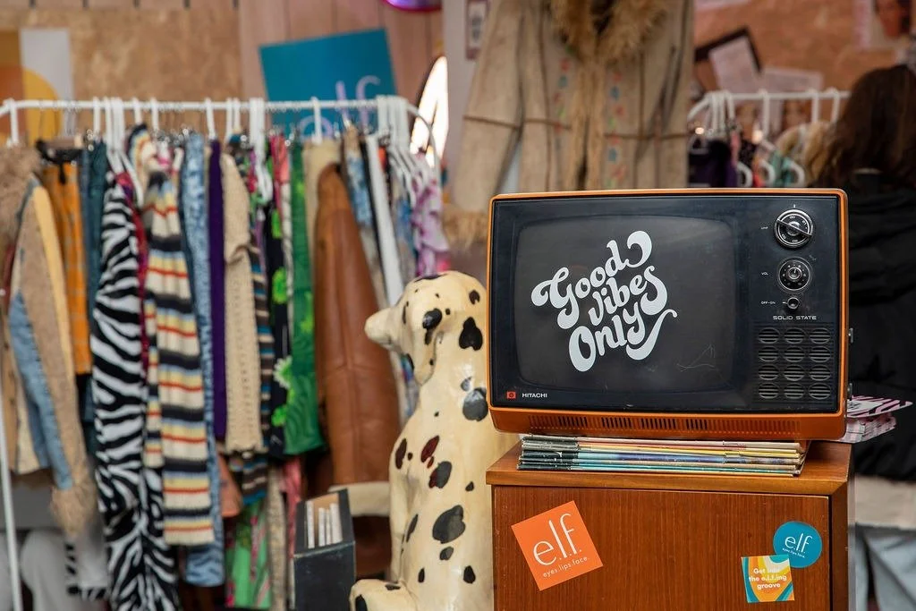Vintage orange portable television with the words "Good Vibes Only" on the screen, a ceramic Dalmatian dog statue, and a rack of colorful clothing in the background.