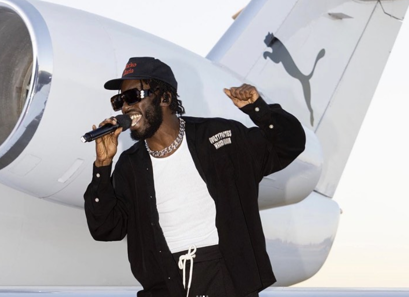 A man with dreadlocks wearing sunglasses, a black cap, a black shirt, and jewelry, holding a microphone and smiling in front of a private jet with the Puma logo.
