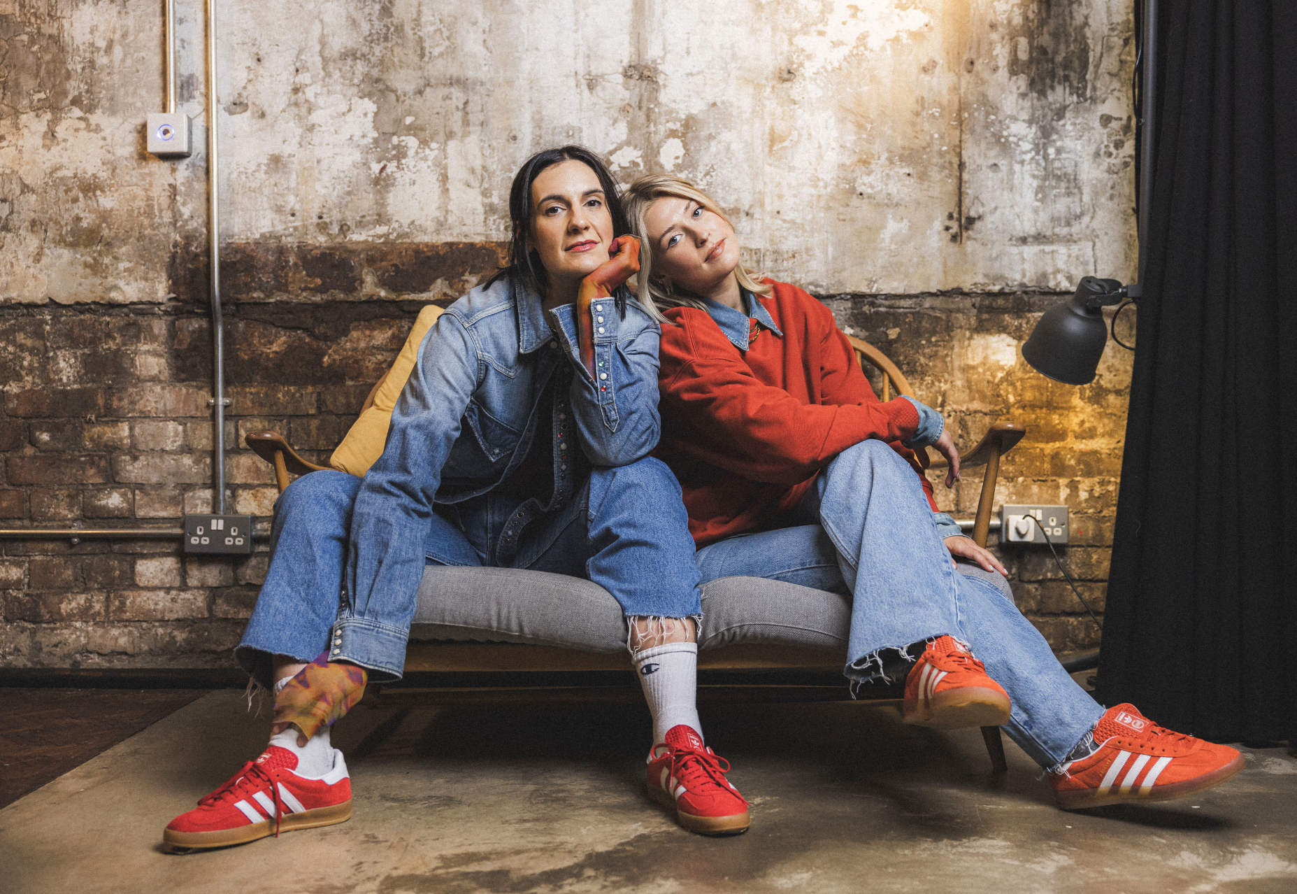 Two women sitting on a gray sofa in a room with exposed brick walls, wearing denim and red jackets, with casual footwear and relaxed poses.