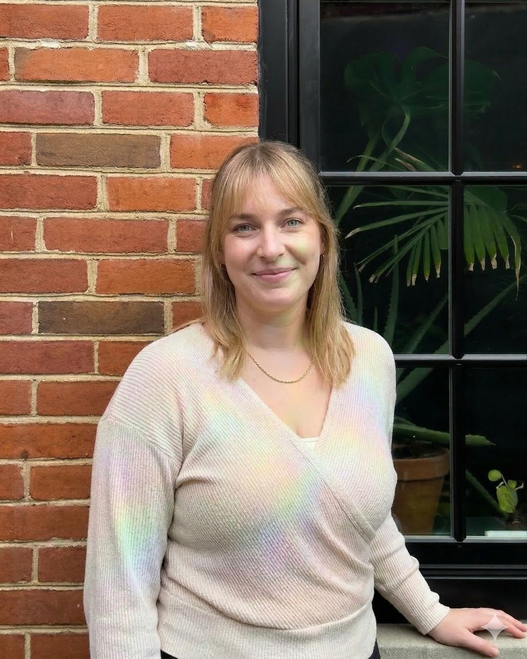 Steph Sobala, Healer at Bath House, smiling and posing outdoors against a brick wall with lush Monstera plants. She is wearing a knit wrap sweater.