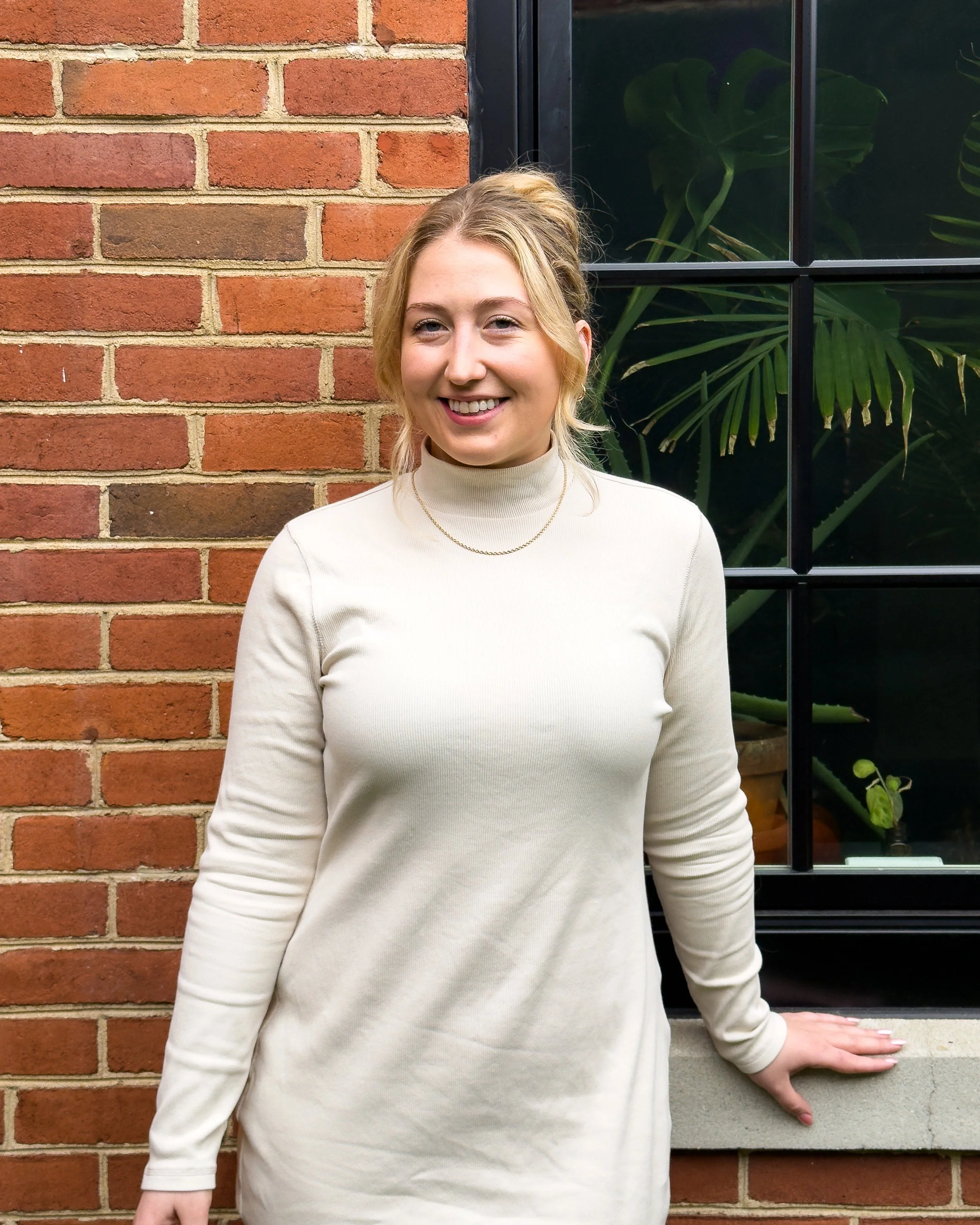 Sam Kelly, Yoga and Movement Healer at Bath House, smiling outdoors in a white dress against a brick wall.