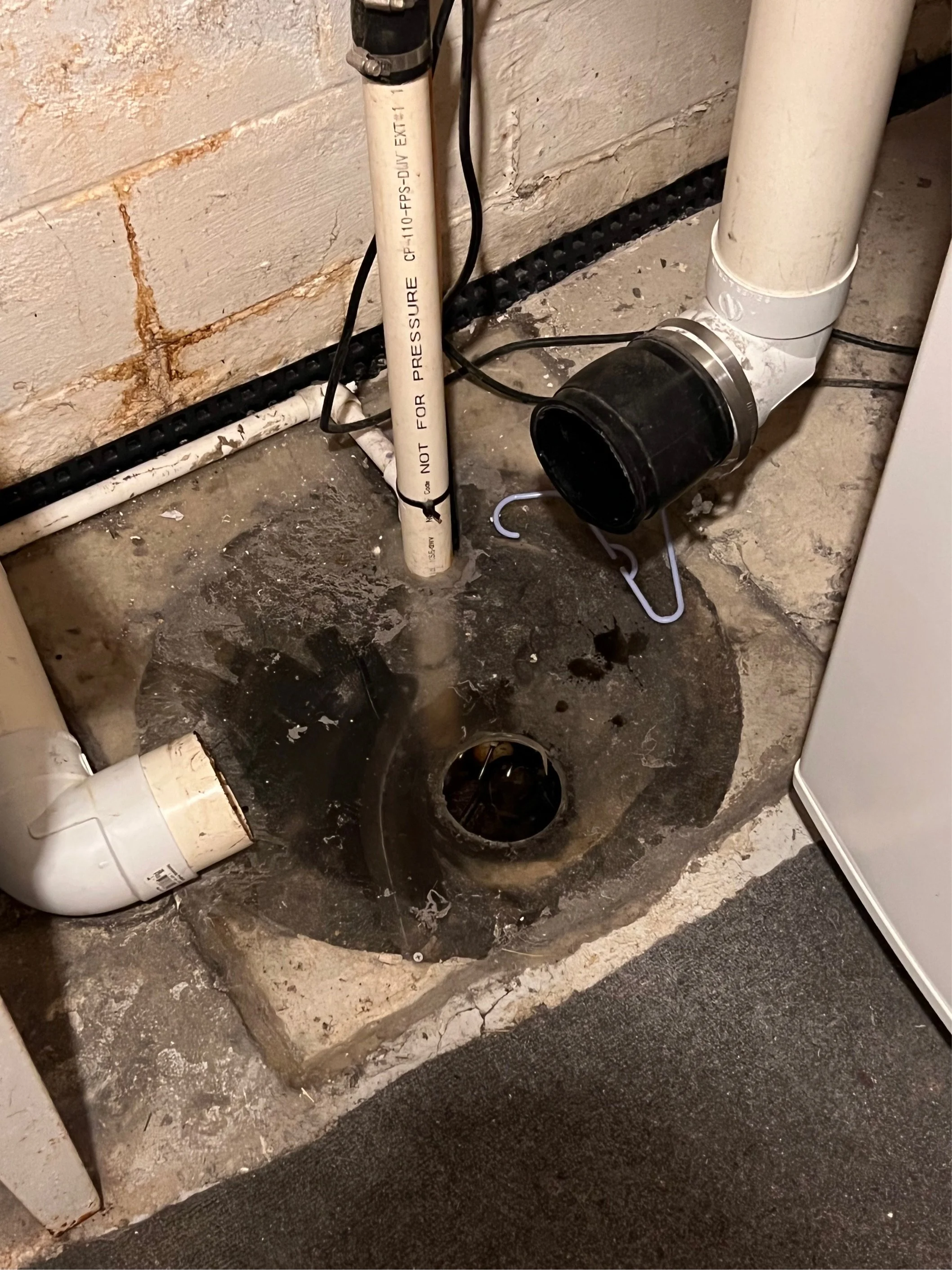 A plumbing drain opening in a basement floor with nearby pipes and an exposed sump pump pipe.