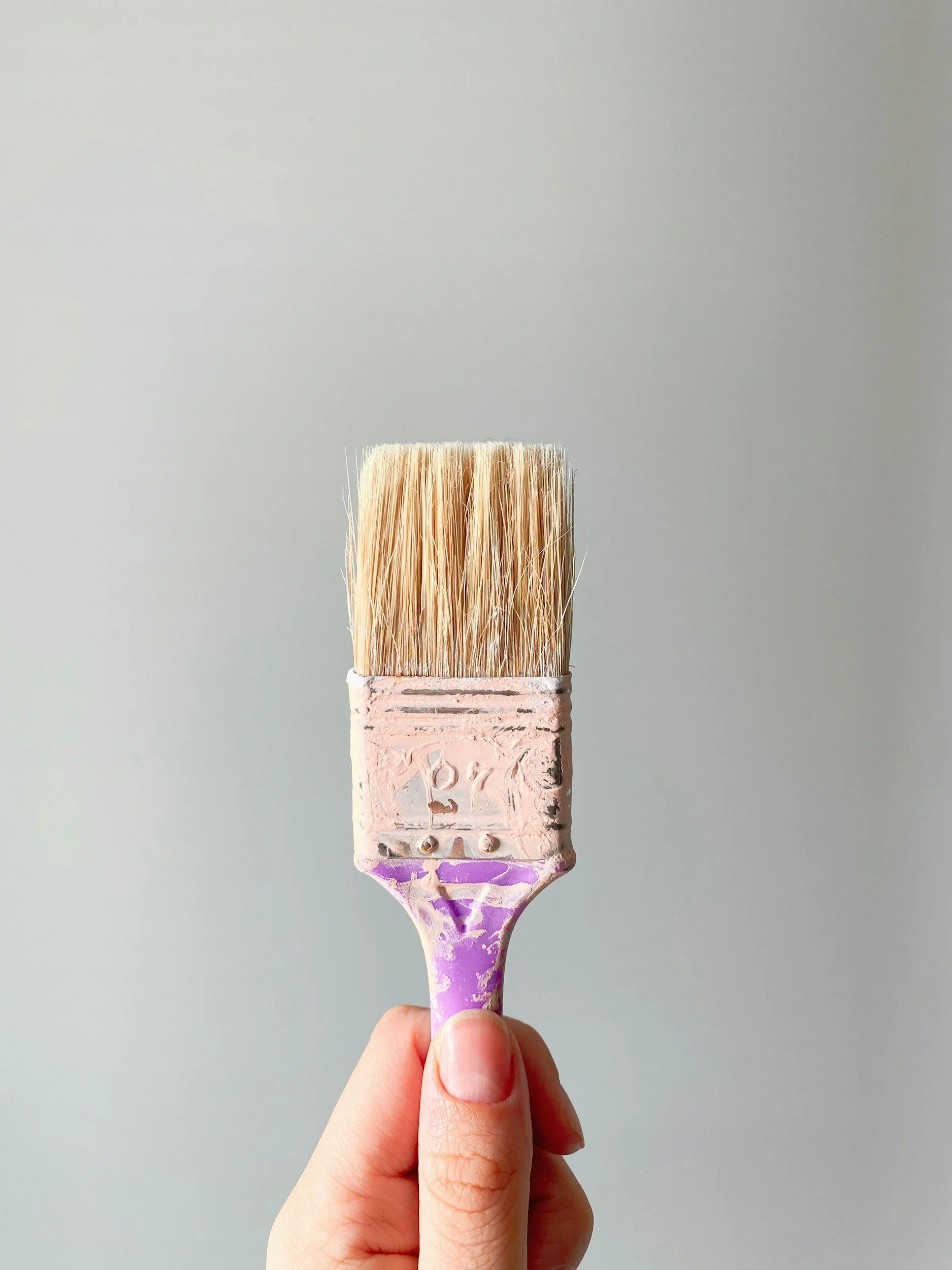 A paintbrush held by a person's hand, with a light gray background.